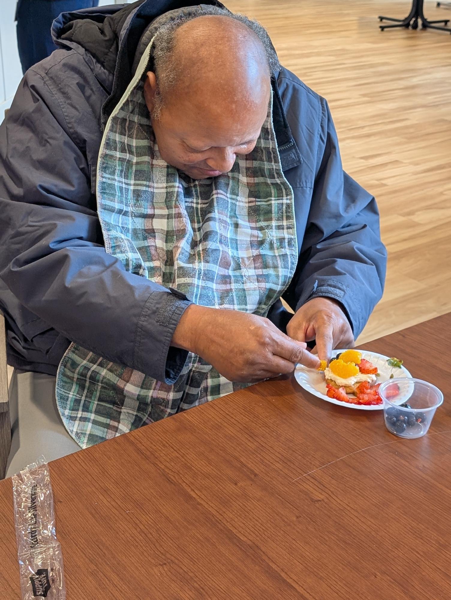 An elderly man with a bald head and gray hair on the sides is wearing a dark jacket and a colorful plaid apron. He is sitting at a wooden table, carefully arranging slices of fruit, such as strawberries and blueberries, on a white plate with a layer 