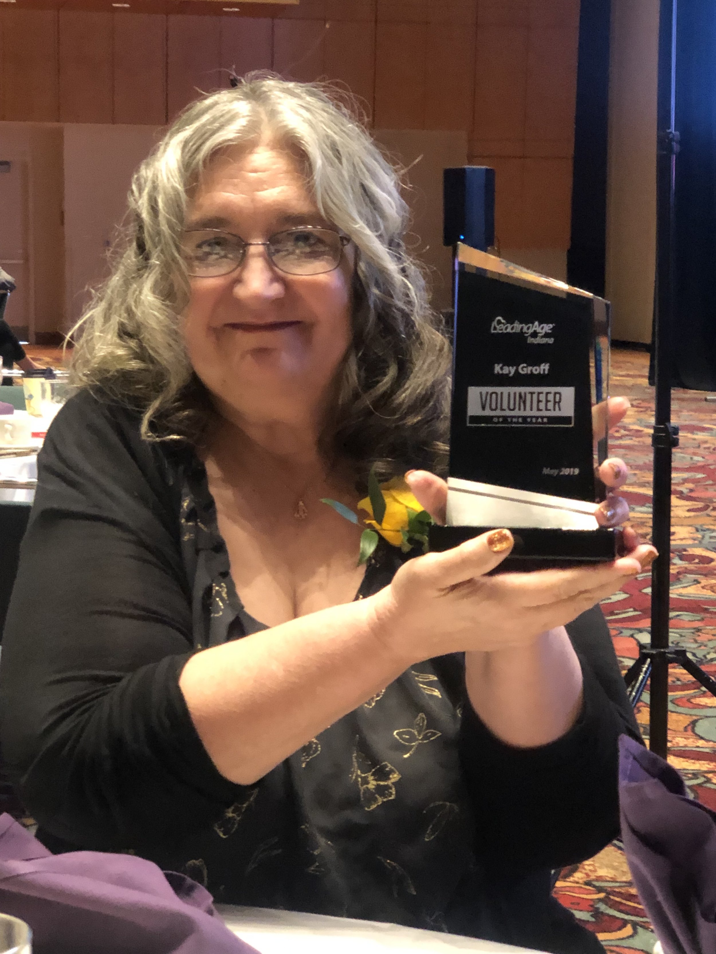 A woman with glasses and curly gray hair holding a trophy labeled 'Leaderage International Kay Groff Volunteer of the Year May 2019' at an indoor event.