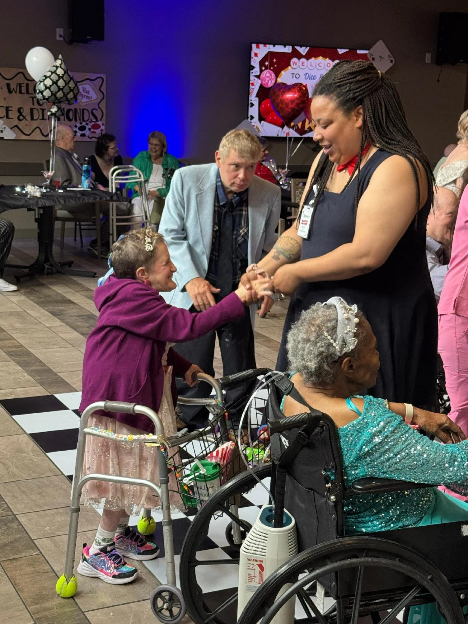 A group of people, including elderly women in wheelchairs and a young girl, are at a celebration event. The young girl is holding hands with a woman, and other elderly people are sitting at tables in the background. There are balloons and a screen wi