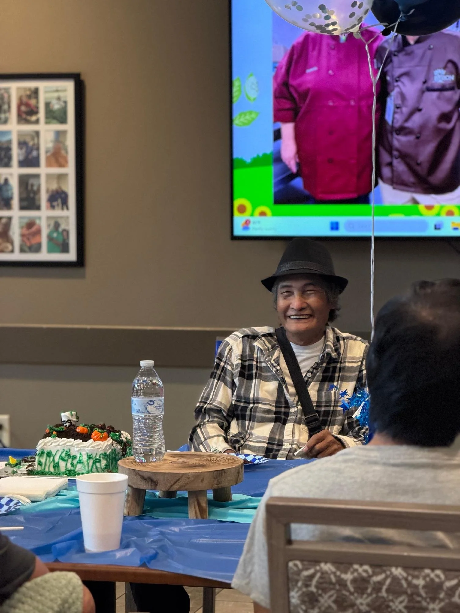 A man wearing a black hat and plaid shirt sits at a table with a decorated birthday cake, a water bottle, and a paper cup. There are balloons and people in the background, indicating a celebration.
