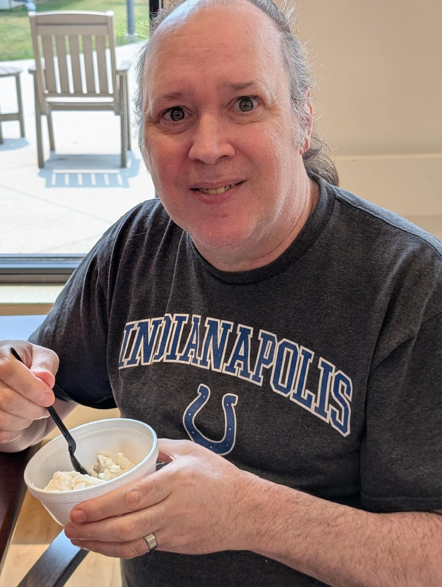 A man with long hair and wearing a gray Indiana Polics shirt is eating ice cream from a white cup with a black spoon at a restaurant table near a window, with outdoor chairs and greenery visible outside.