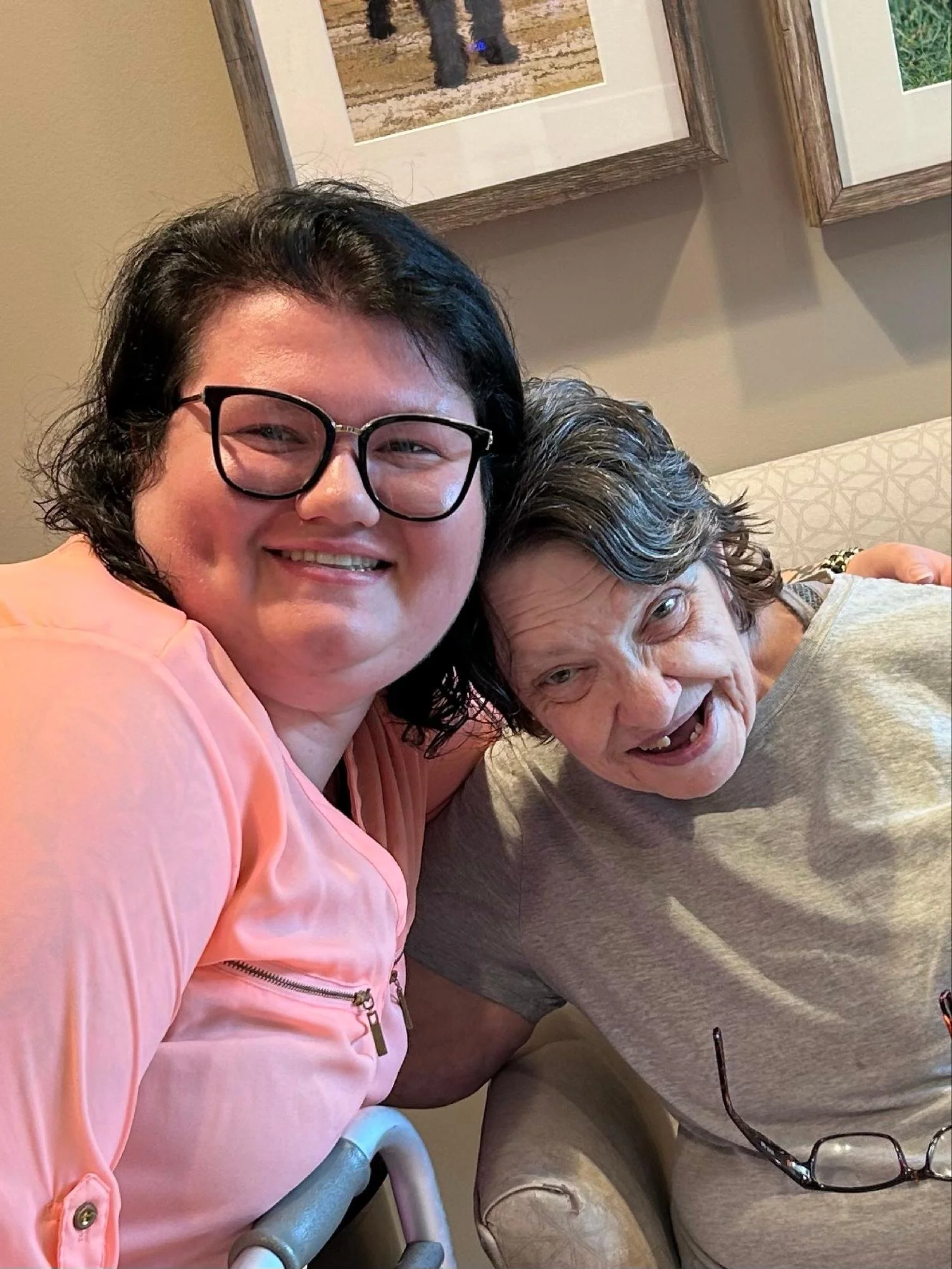 Two women smiling for a selfie in an indoor setting. The woman on the left has black hair, glasses, and is wearing a pink jacket. The woman on the right has gray hair and is wearing a gray shirt. There are framed pictures on the beige wall behind the