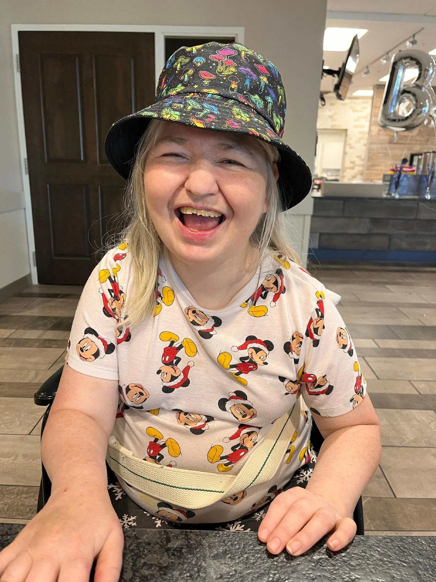 A woman wearing a Mickey Mouse patterned shirt, a bucket hat with colorful mushroom prints, and a white belt, smiling and sitting at a table in a restaurant or cafe.
