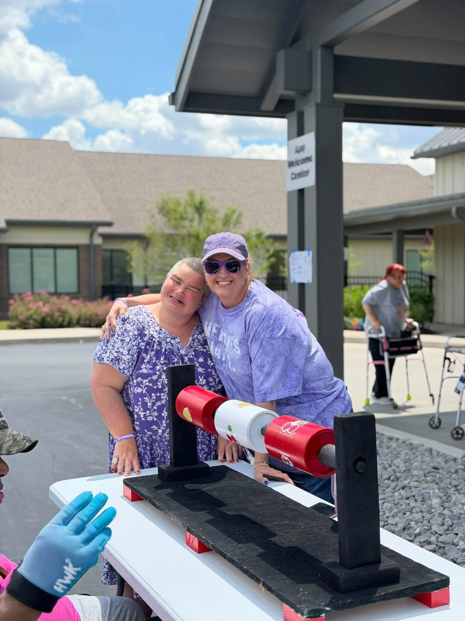 Two women smiling and hugging at a science demonstration table with a rocket model, with a person in the background wearing sunglasses and a gray jacket, and another person with red hair and a walker in the distance.