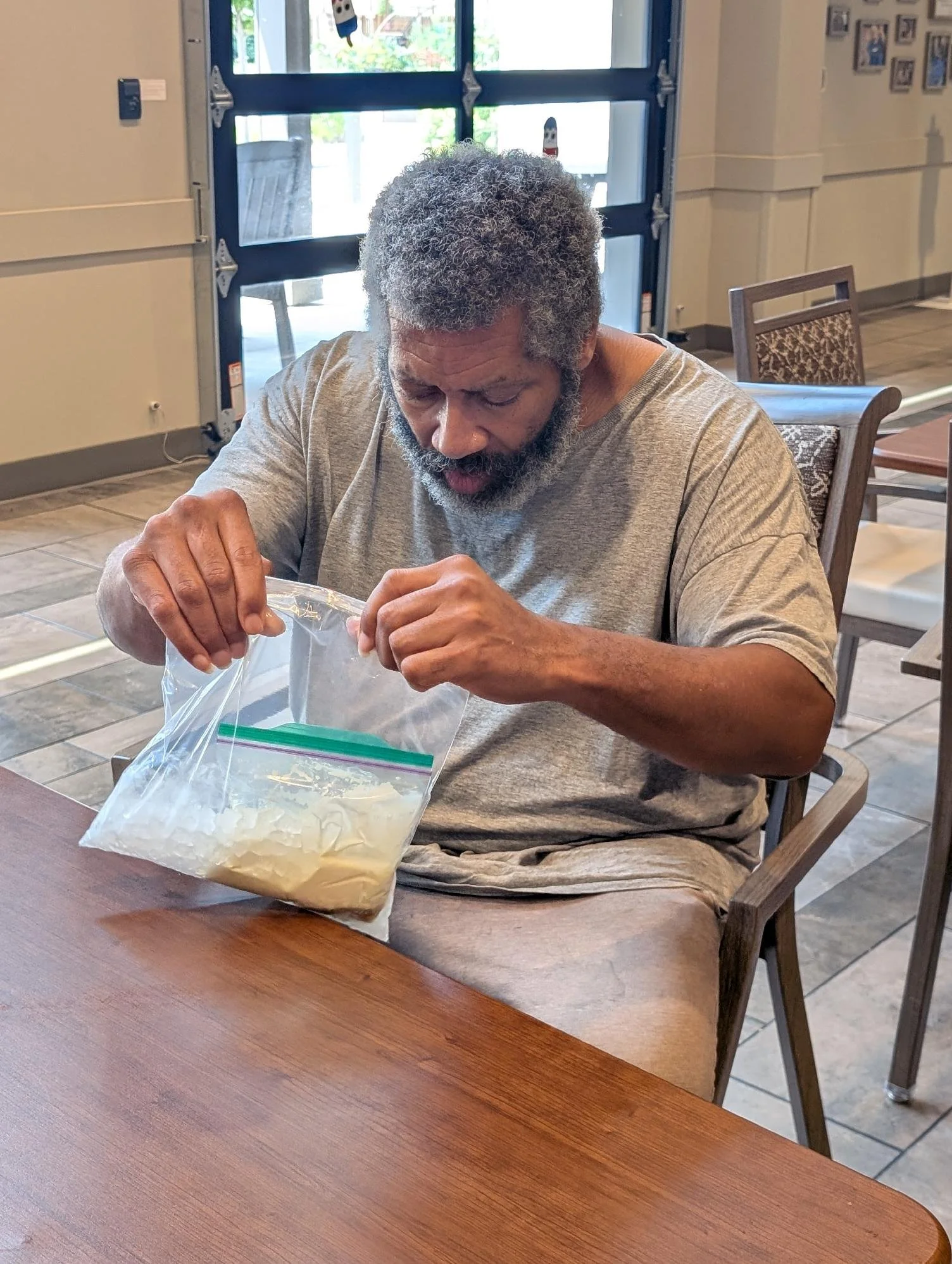 An older man with gray curly hair and a beard sitting at a wooden table, looking down and opening a plastic bag with a yellowish substance inside.