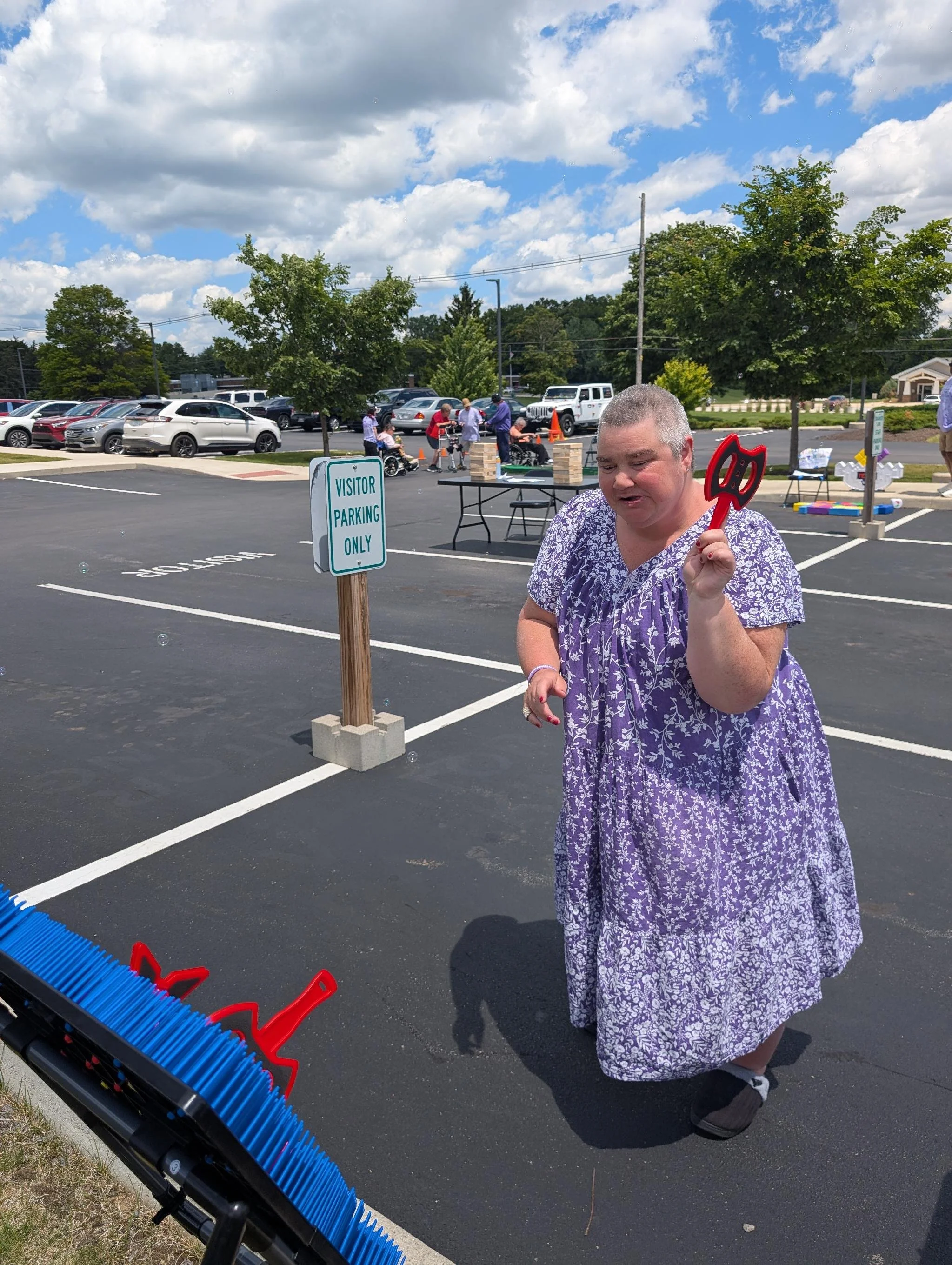 A woman in a purple floral dress holding a red spatula stands in a parking lot next to a 'Visitor Parking Only' sign during daytime. In the background, a group of people, some in wheelchairs, are playing Jenga at a table. Several trees and parked car
