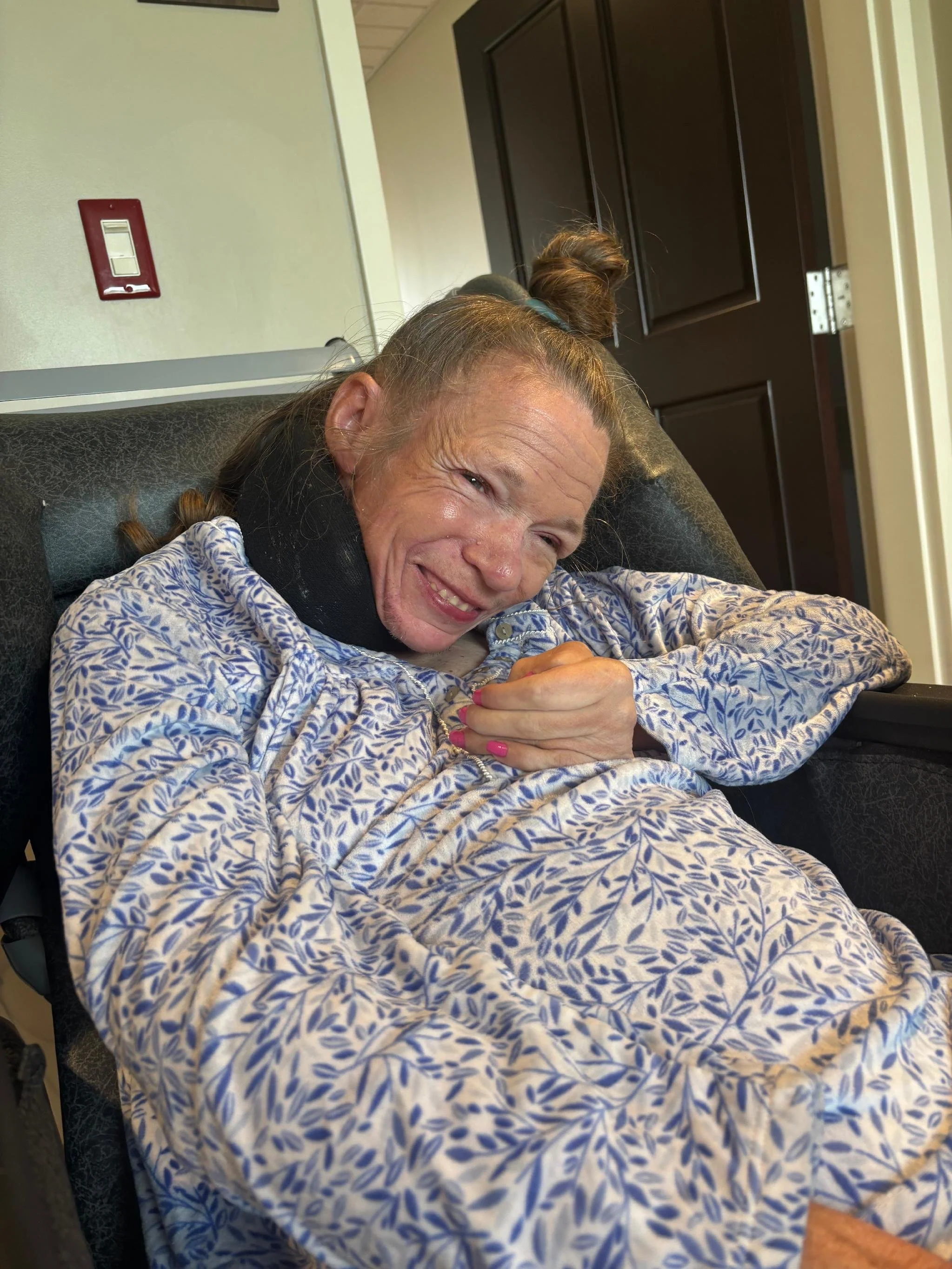 A woman with braided hair and a smile resting on a couch, wearing a floral patterned shirt, with a dark door and light-colored wall in the background.