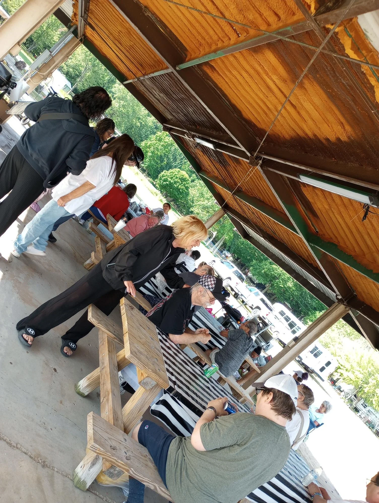 People gathered around a long outdoor table with a striped tablecloth, under a canopy, at an outdoor market or fair, with cars and green trees in the background.