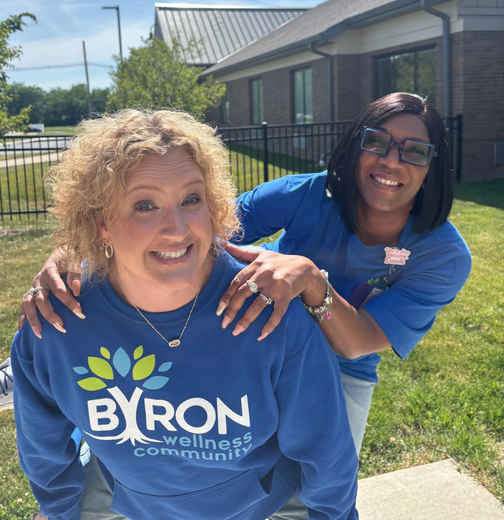 Two women smiling outdoors, one with blonde curly hair and the other with black straight hair and glasses, wearing blue shirts with a logo and text, standing in front of a house and a black fence, sunny day.