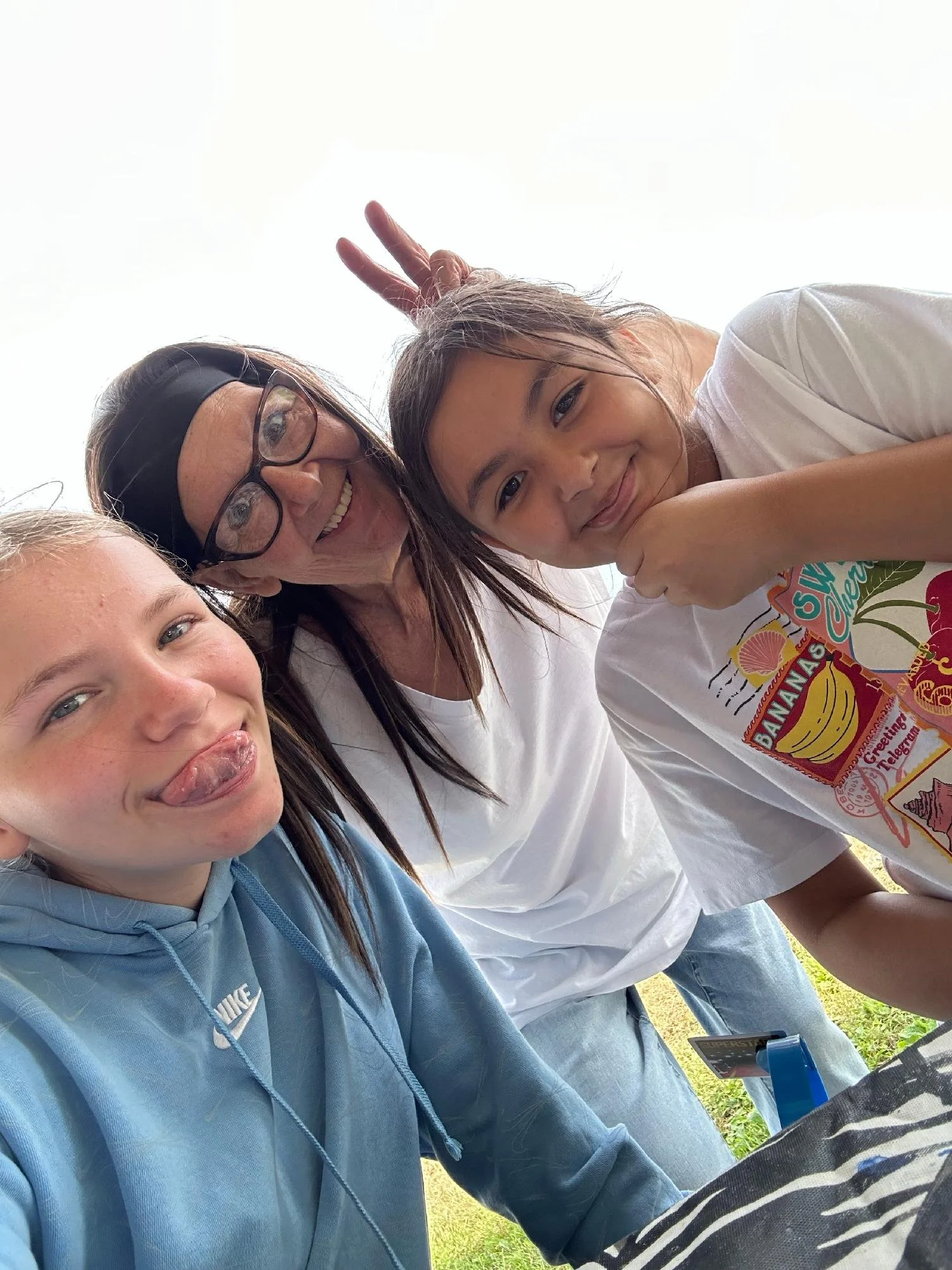 Three women outdoors, taking a selfie. The woman on the left has blonde hair, the woman in the middle has brown hair, and the woman on the right has brown hair and is making a peace sign behind her head. All are smiling.