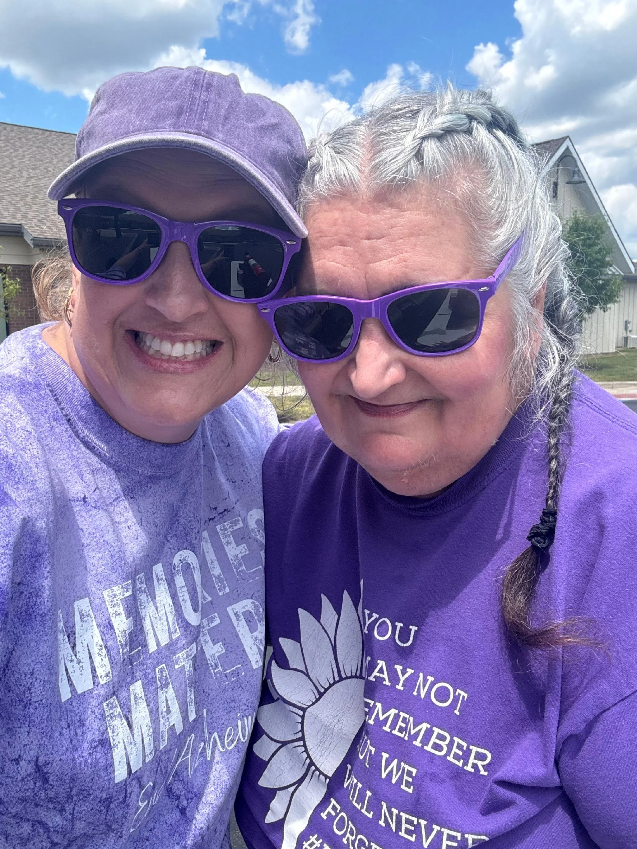 Two women smiling close to each other outdoors on a sunny day, both wearing purple sunglasses and purple shirts, with houses and blue sky with clouds in the background.