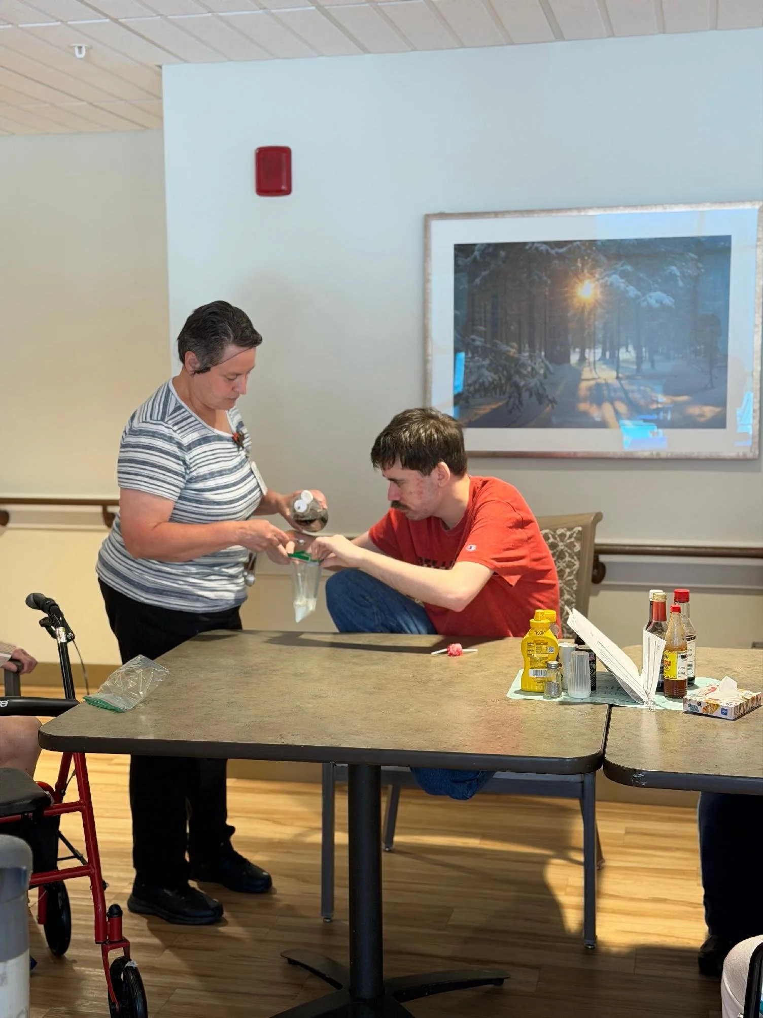 A woman in a striped shirt handing medication to a young man in a red shirt at a table with condiments and food supplies, in a room with light-colored walls and wooden flooring, and a large framed picture of a snowy landscape hanging on the wall.