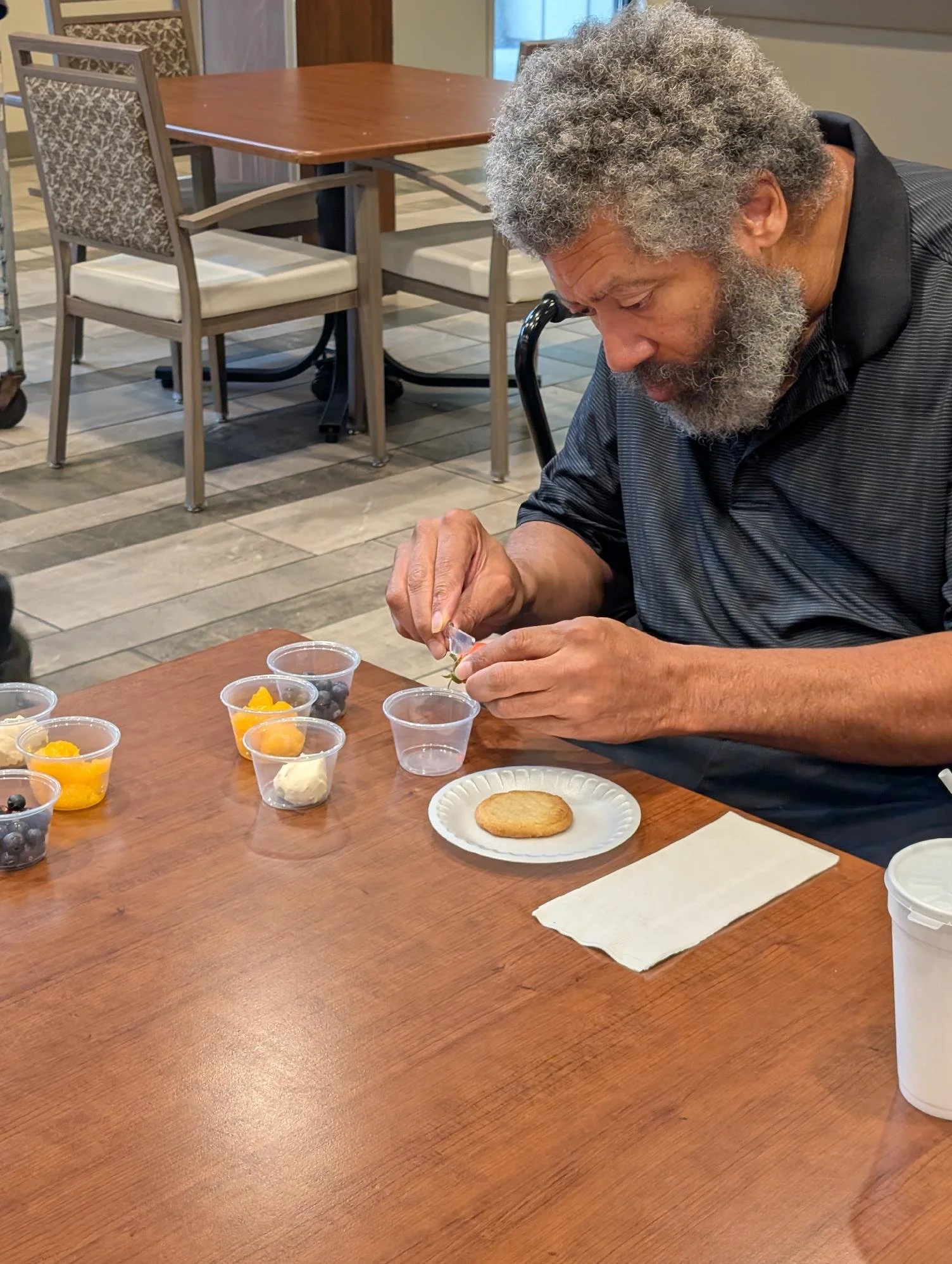 An elderly man with gray curly hair and beard sitting at a wooden table, looking at a small vegetable on his hand, with various cups of colorful fruit and a cookie in front of him in a restaurant or cafe setting.