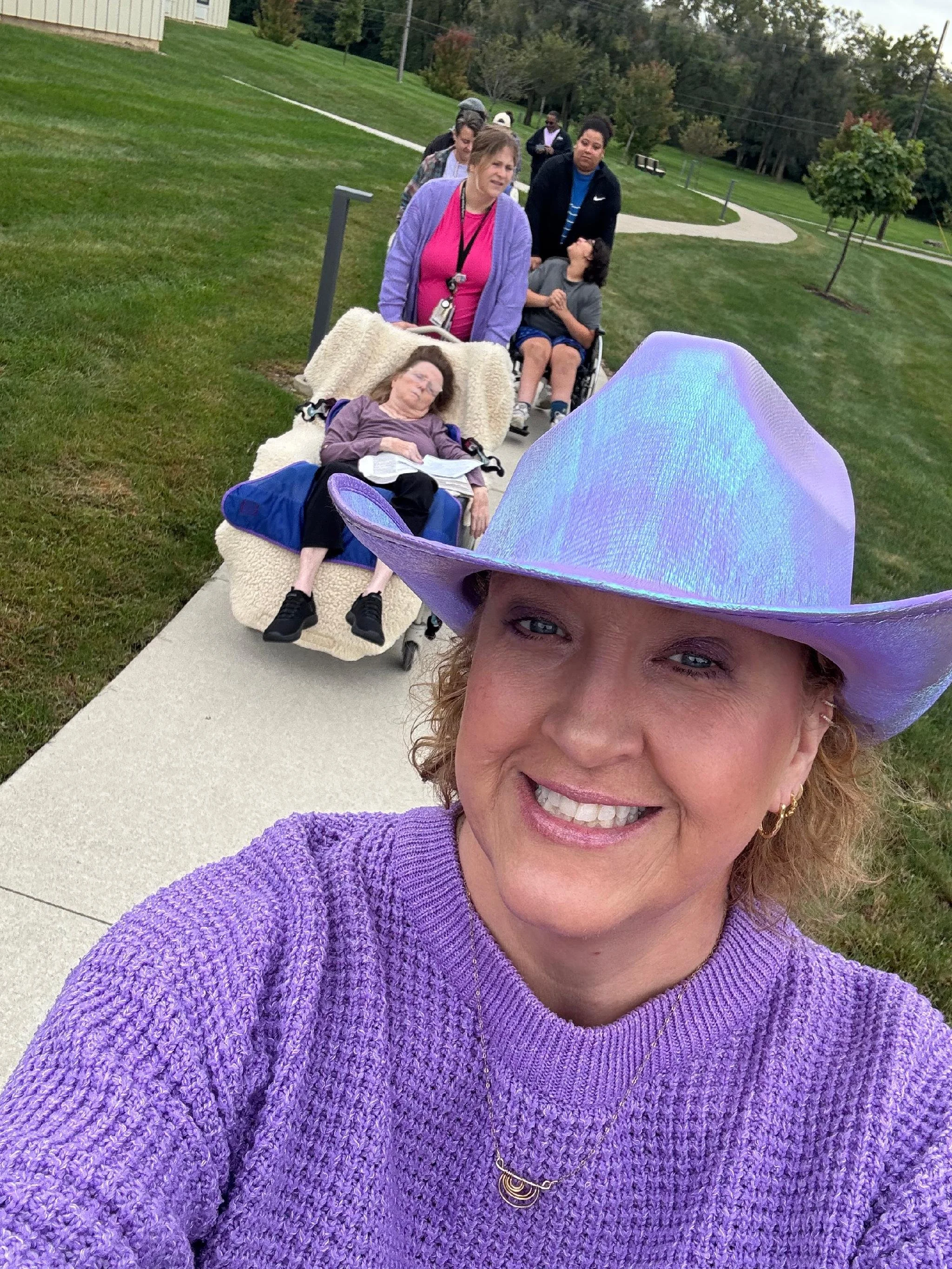 A woman wearing a purple sweater and a large purple cowboy hat, smiling at the camera, outdoors on a sidewalk with a group of people walking behind her on a grassy area.