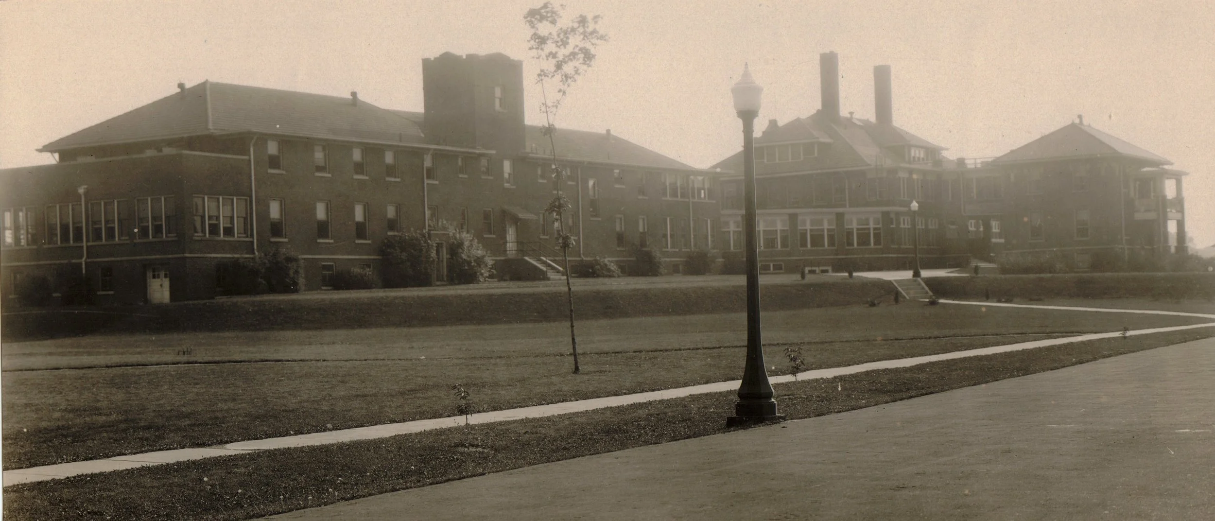 Old black and white photograph of large multi-story brick buildings with many windows, set in a grassy area with sidewalks and vintage street lamps.