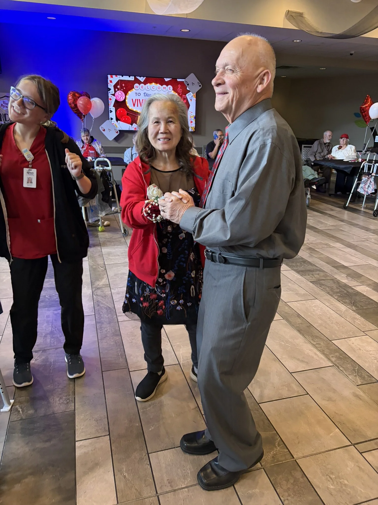 A senior man in a gray suit holding hands and smiling with a woman in a red jacket at a celebratory event, with birthday themed decorations and other people in the background.