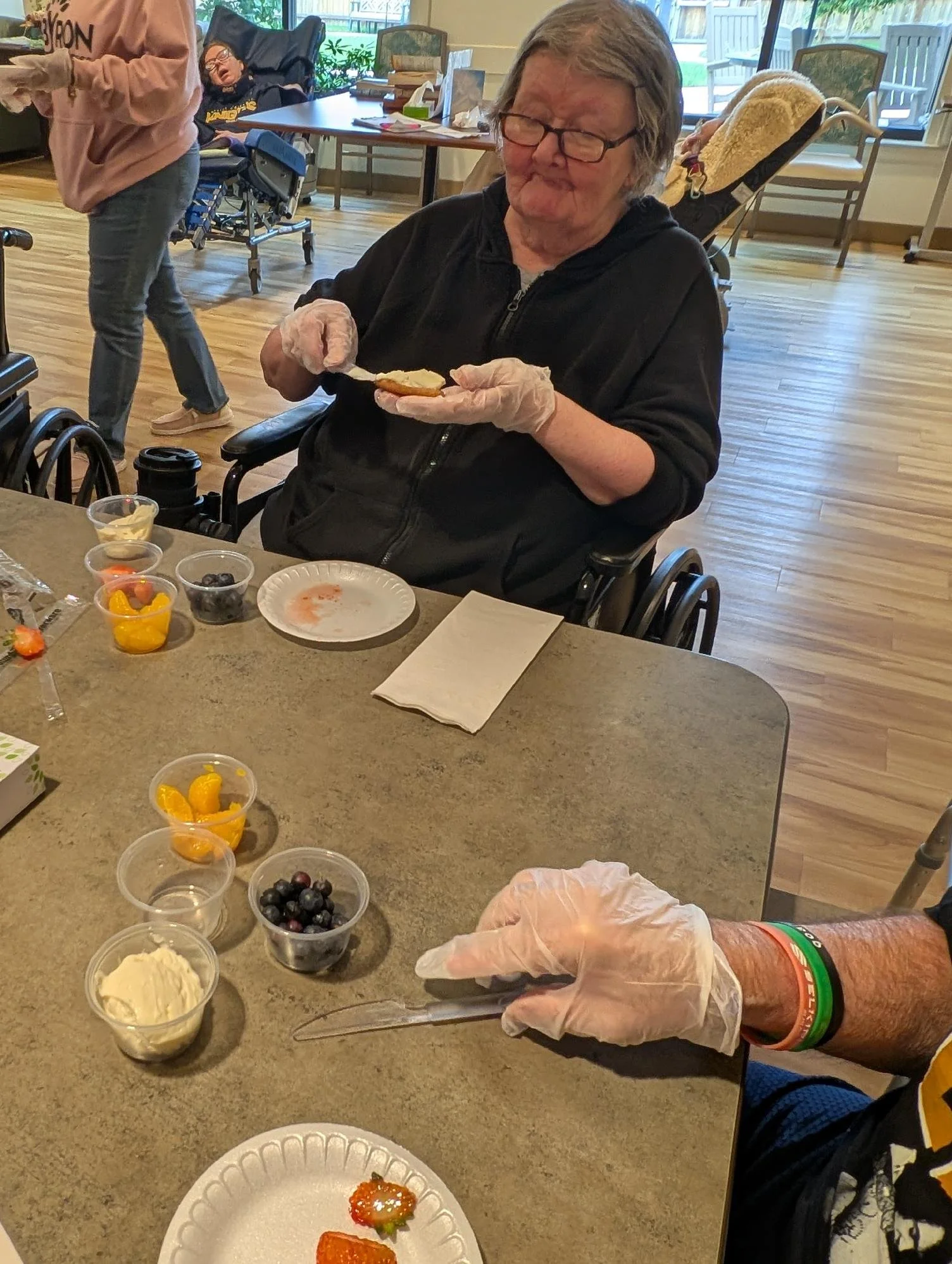 An elderly woman with glasses and gray hair, sitting in a wheelchair, wearing a black hoodie, and spreading frosting on a cookie. The table in front of her has various bowls of fruit, cream, and other desserts. There is a person in a pink hoodie in t