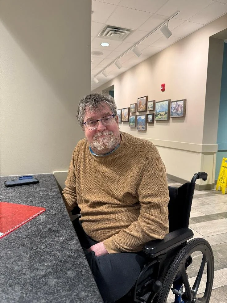 A man with glasses and gray hair sits in a wheelchair at a table in a room with artwork on the wall.