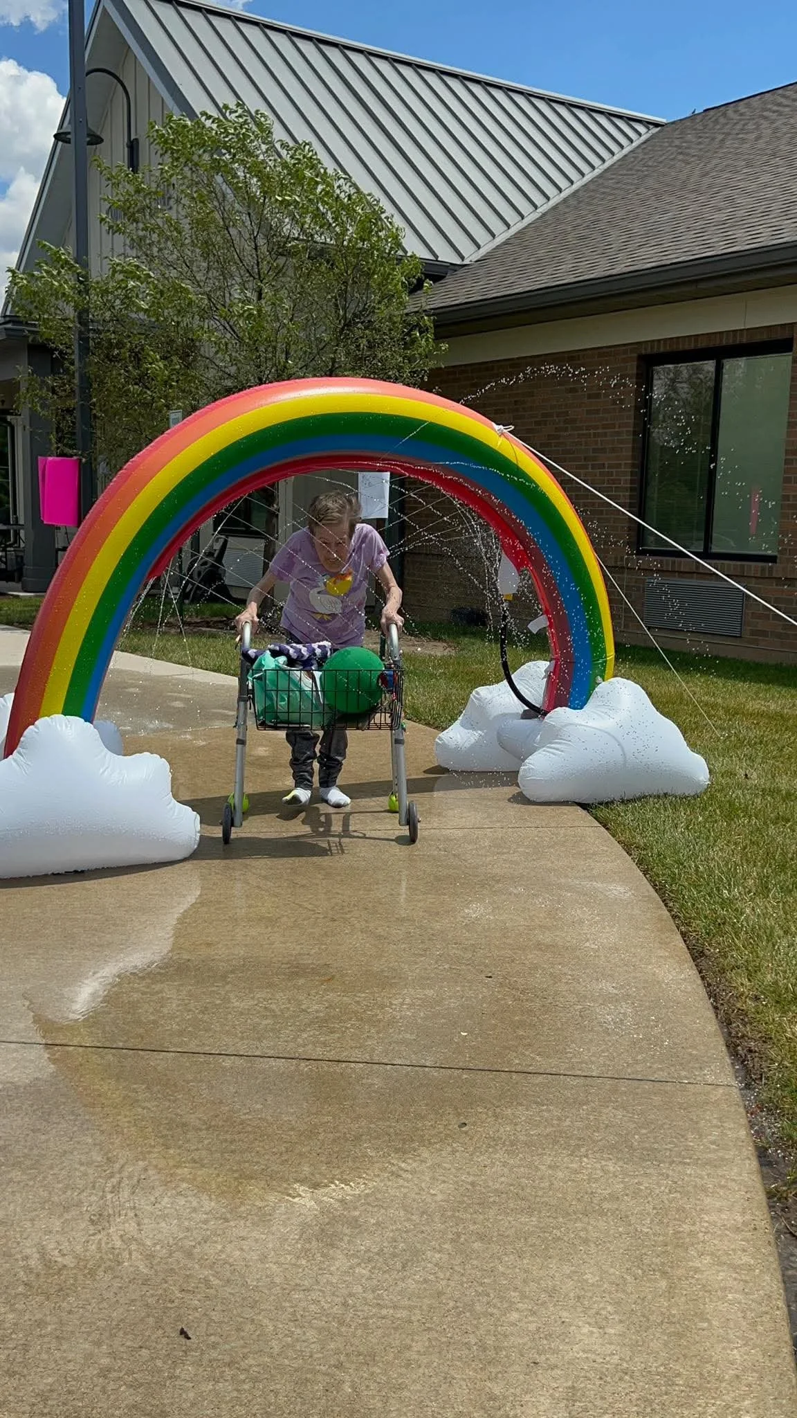 A woman with a walker walking through a water spray beneath an arch rainbow with clouds at each end.
