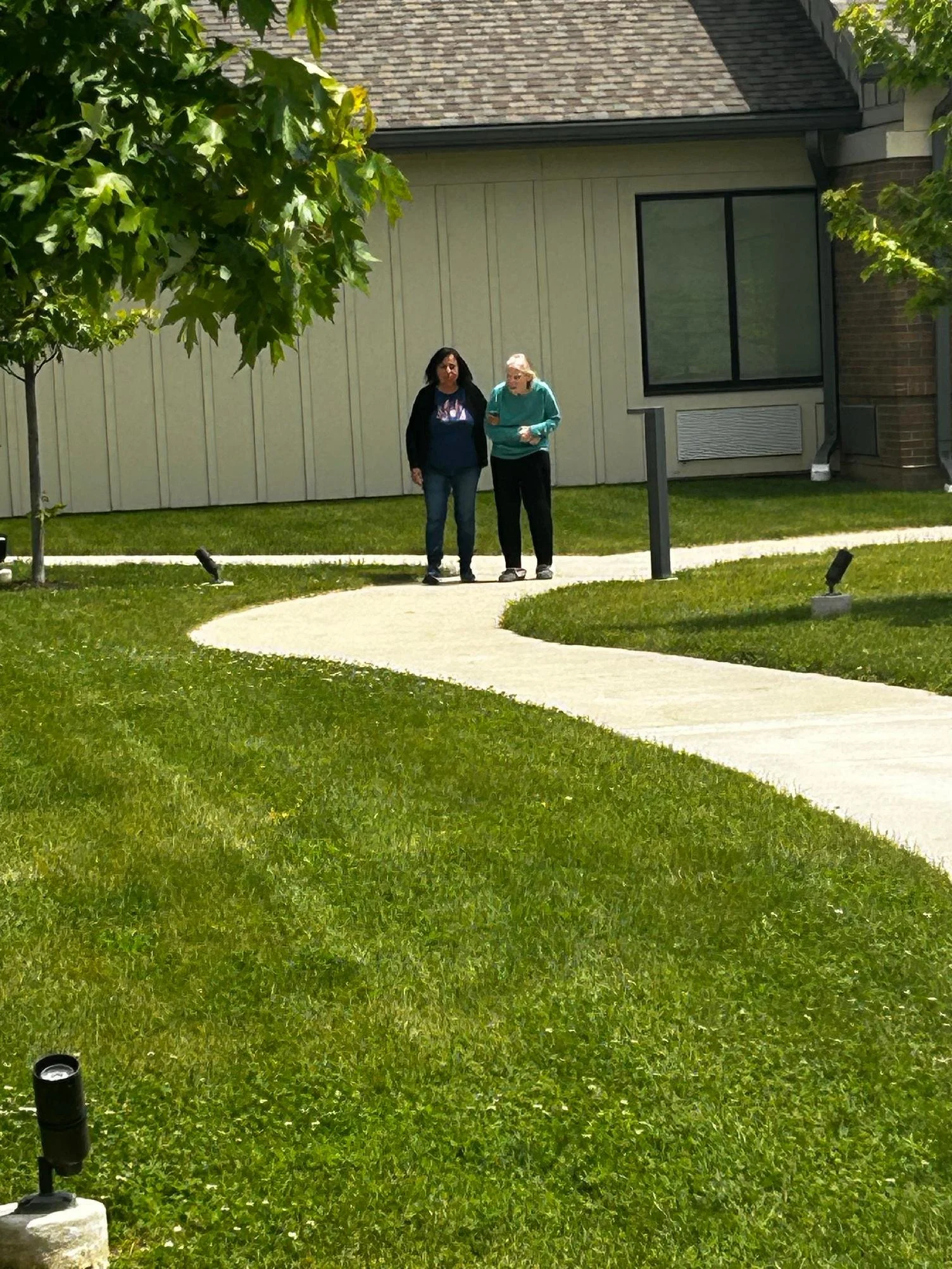 Two women walk along a curved concrete path in a grassy yard in front of a house. The house has cream siding with a window and brick on one side. A tree with green leaves partially frames the scene, and outdoor lighting fixtures are in the grass.