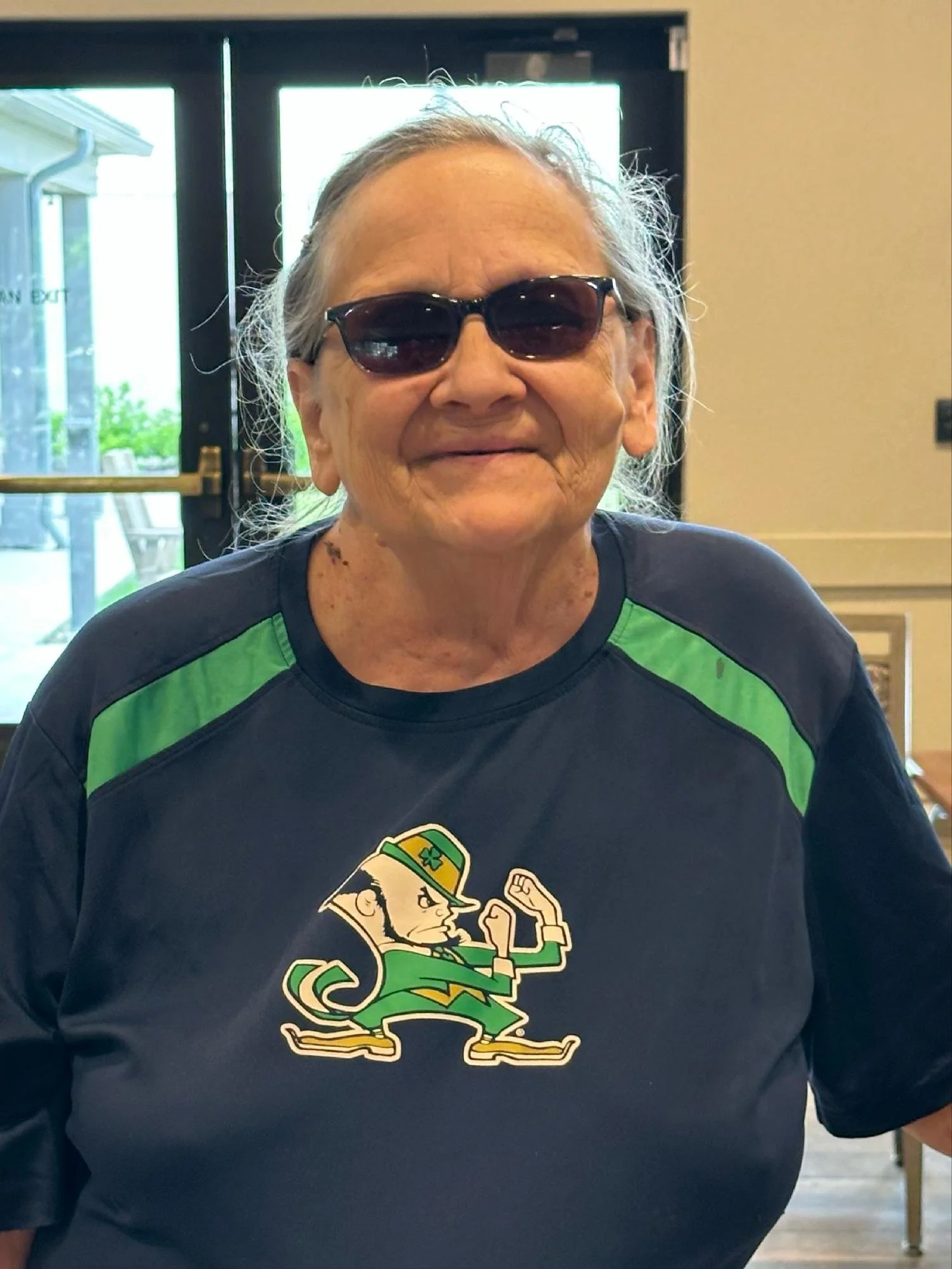 Older woman with gray hair wearing sunglasses and a black and green sports shirt with a cartoon cartoon of a person with a hat on it.