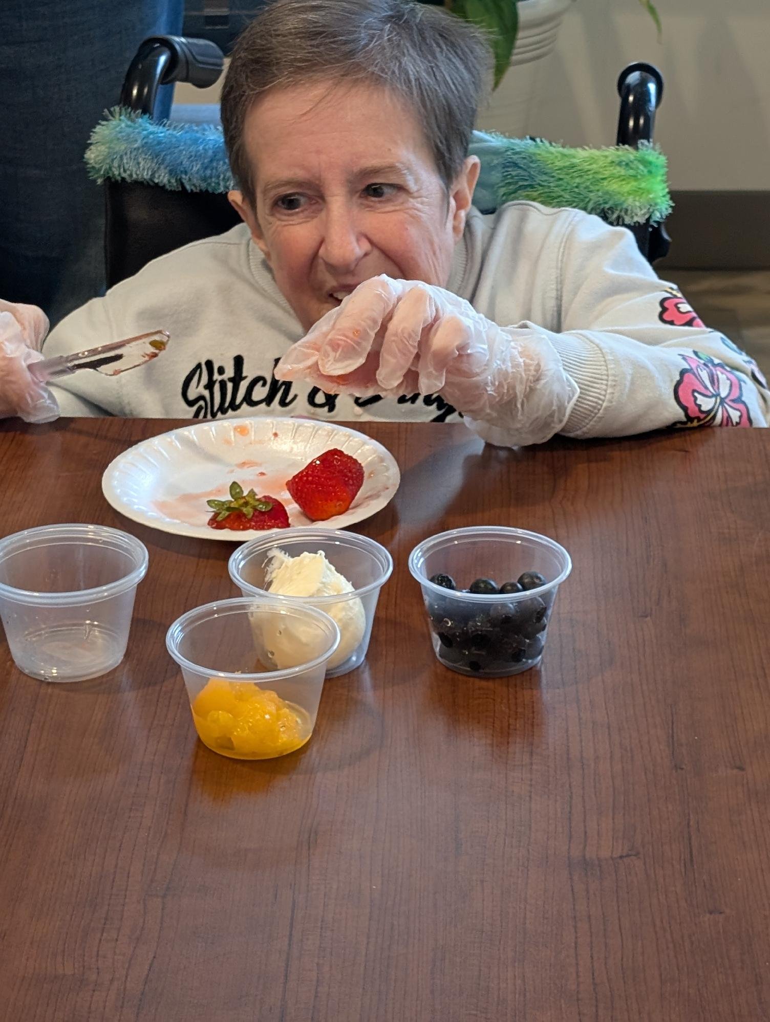 An elderly woman with short brown hair wearing a white sweatshirt with pink floral patches on the sleeves, sitting in a wheelchair, reaching for a strawberry on a paper plate in front of her. There are small containers of various desserts and fruits 