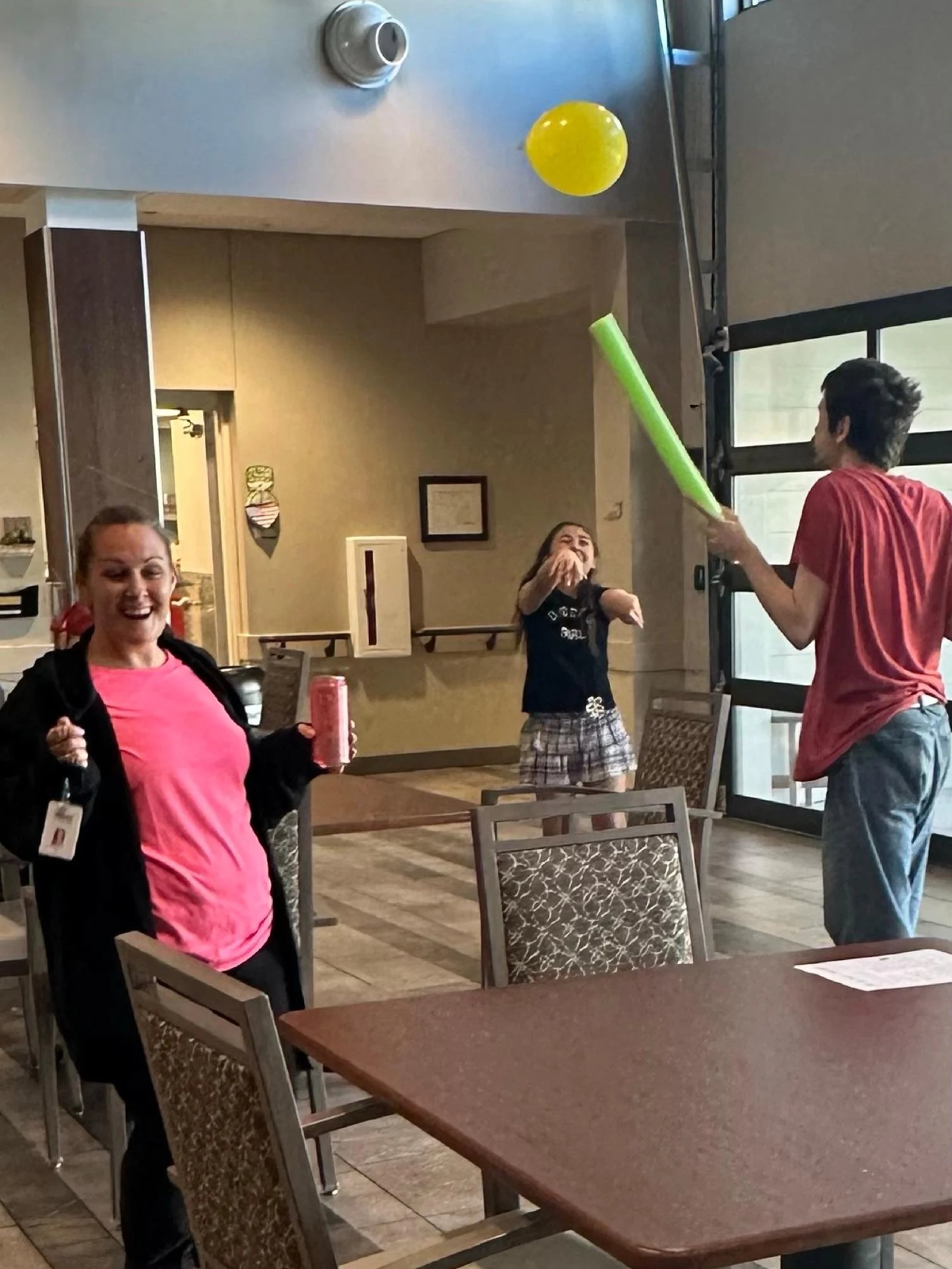 People in a restaurant playing with balloons. One person is holding a green balloon with a stick, aiming at a yellow balloon in the air. Two women are laughing, one holding a red soda can.