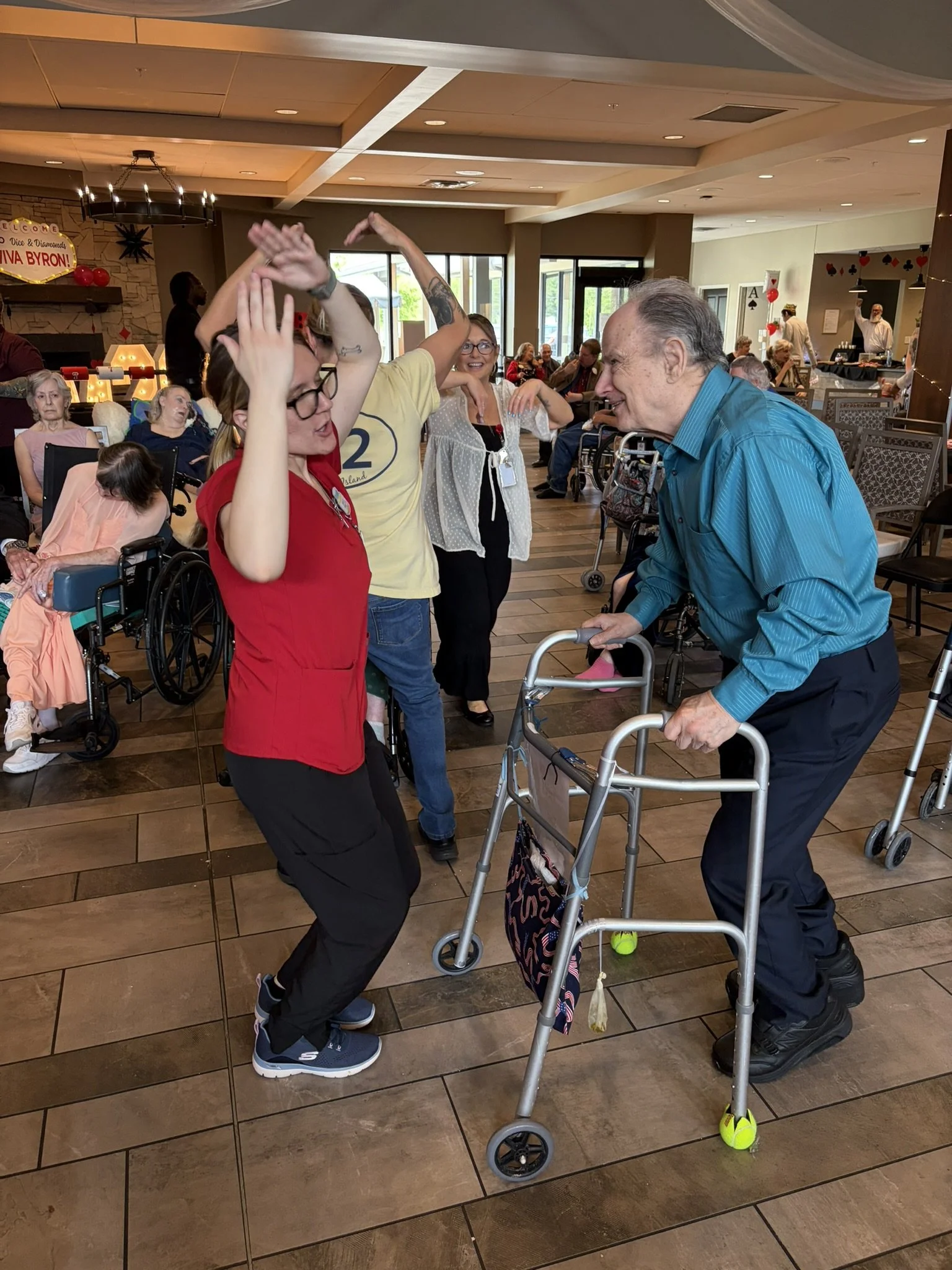 Multiple elderly and disabled people in wheelchairs and staff members dancing and celebrating in a dining area decorated with balloons and cards, likely for a special occasion.