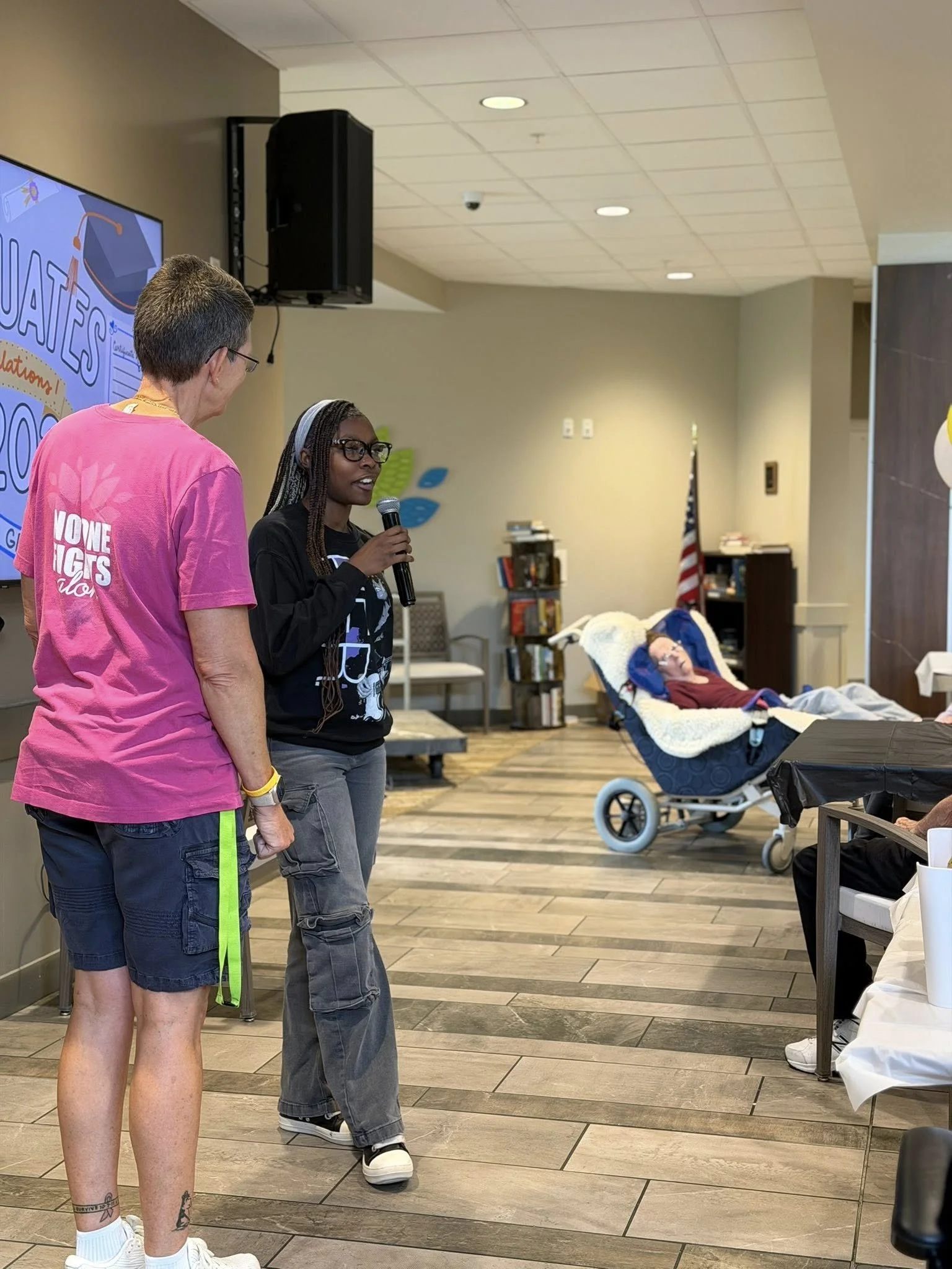 Two women are standing and talking in a room with a pediatric patient in a hospital bed nearby. The woman on the left has short hair and tattoos on her lower legs, and is wearing a pink T-shirt and shorts. The woman on the right is holding a micropho