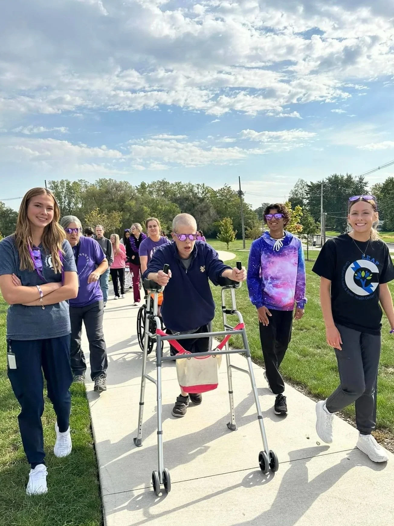 Group of diverse young women and a person in a wheelchair walking outdoors on a sidewalk, smiling, during a sunny day with a partly cloudy sky and trees in the background.