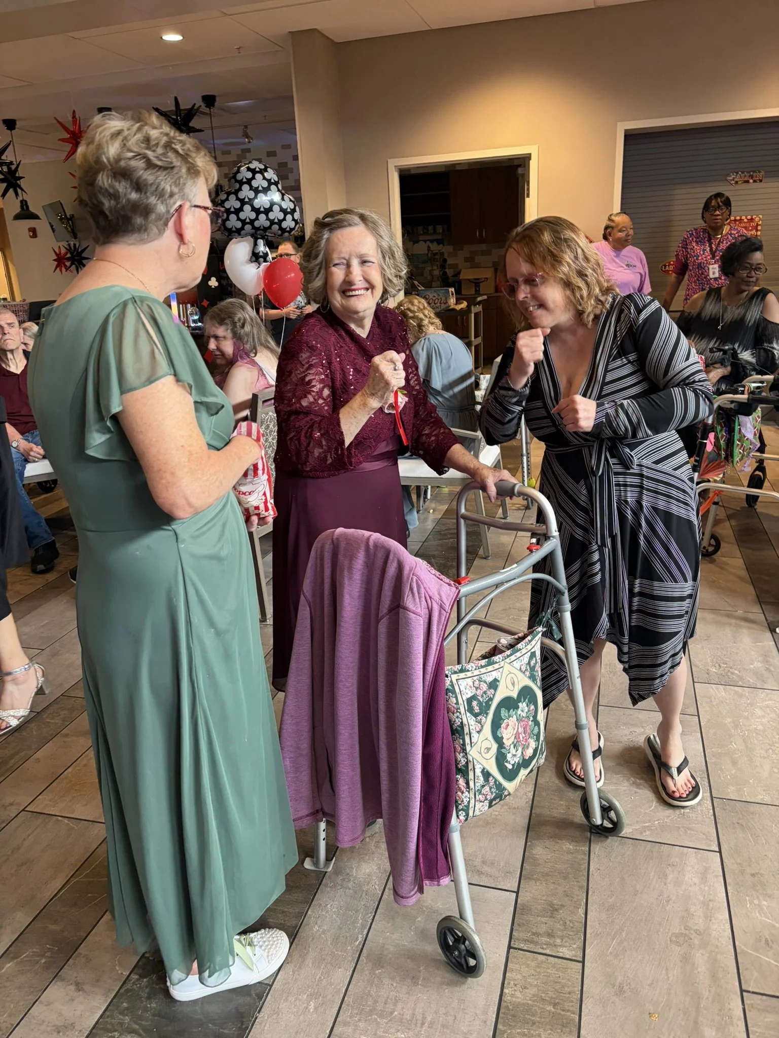 Three women are talking and laughing at a social gathering, with a walker in front of them and balloons in the background.