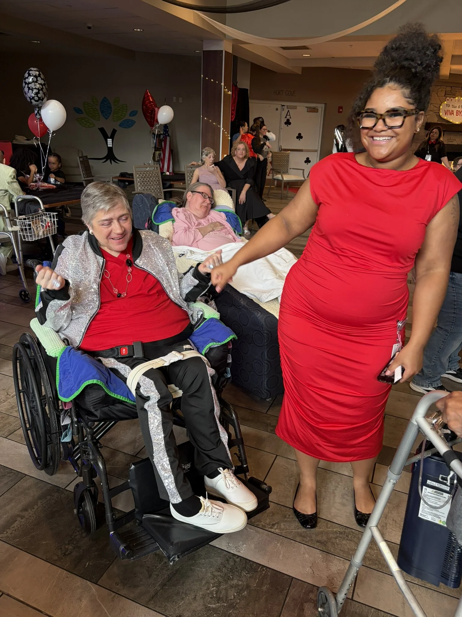 Women wearing red dresses interacting at a social gathering in an indoor event space decorated with balloons and American flags.