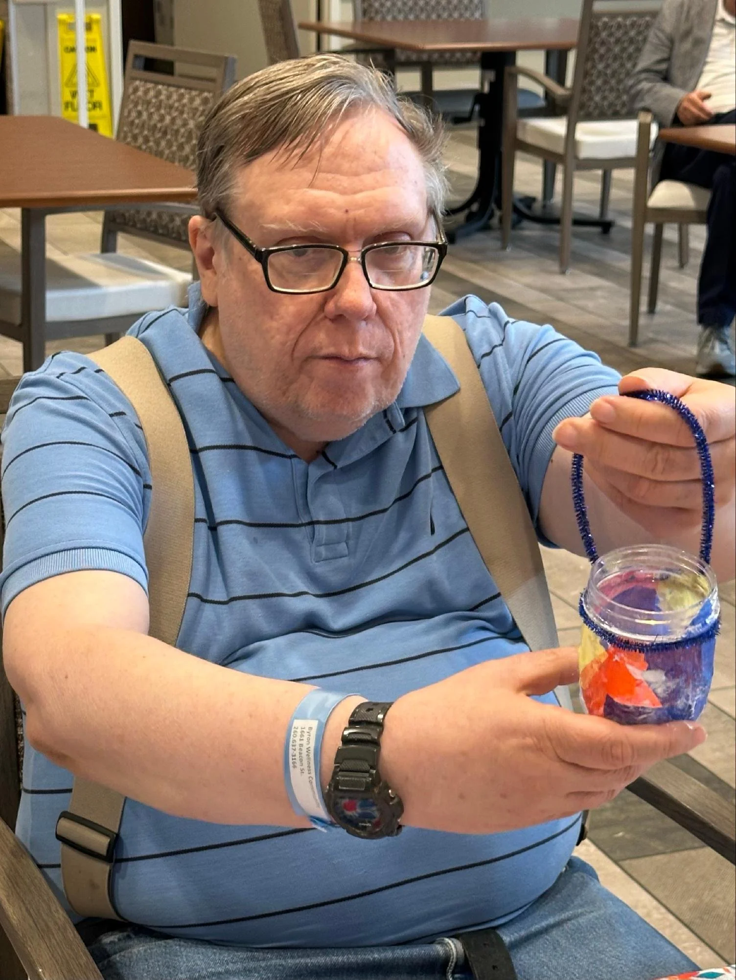 A middle-aged man with glasses and gray hair sitting in a restaurant, holding a colorful beverage with a straw. He is wearing a blue striped shirt, a watch, and a medical wristband. There are tables and chairs in the background, and a caution sign is
