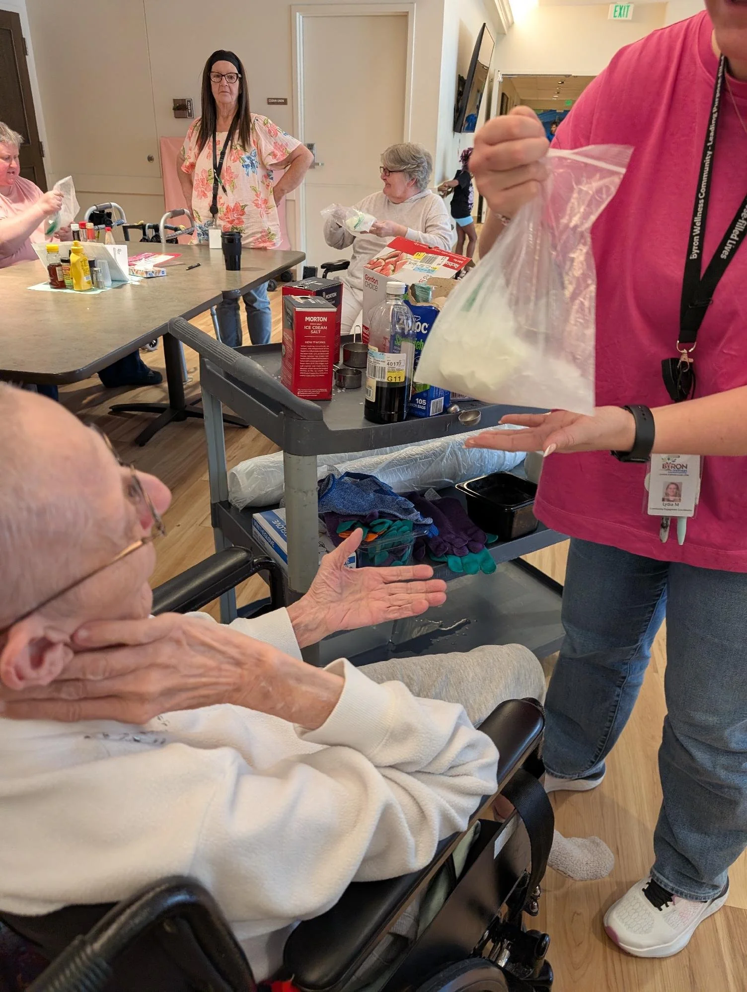 An elderly woman sitting in a wheelchair extends her hand towards a woman wearing a pink shirt who is holding a plastic bag with items. Several other people are around, some sitting at a table with food and condiments, in an indoor setting with woode