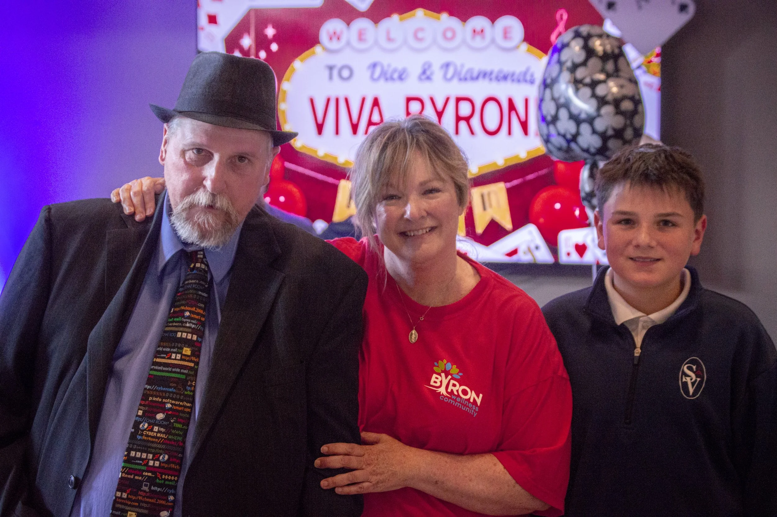 Three people standing in front of a red and white sign that says 'Welcome to Dice & Diamonds Viva Byron.' The man on the left is wearing a dark suit, a colorful tie with computer code design, and a fedora hat. The woman in the middle is wearing a red