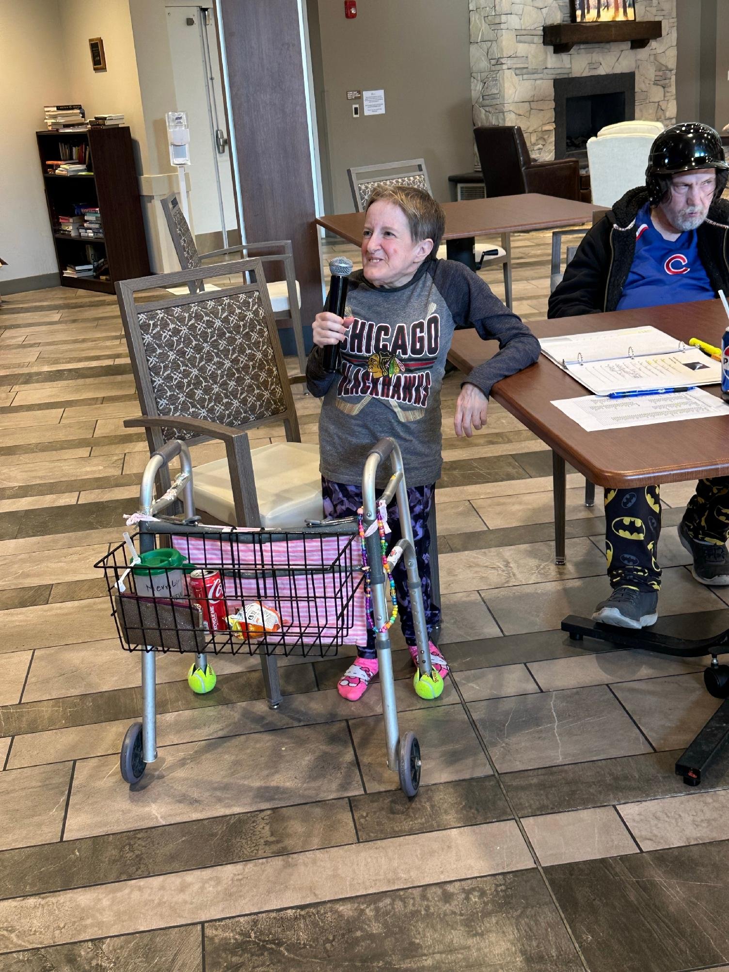 Living room with a woman holding a microphone and a man wearing a motorcycle helmet sitting at a table, with a shopping cart of groceries in the foreground.