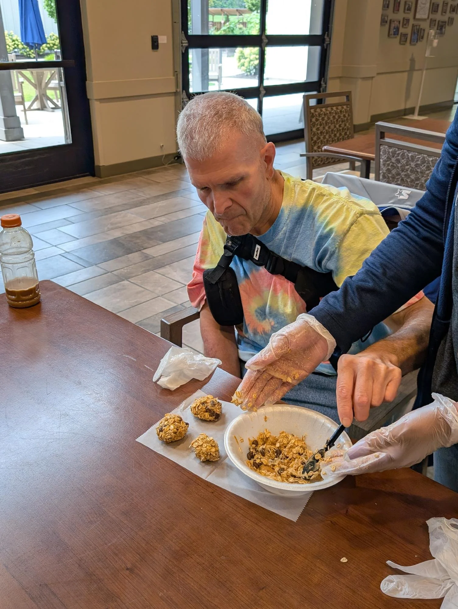 A man with short gray hair sitting at a table, wearing a colorful tie-dye shirt, as someone prepares food nearby. On the table, there are several homemade energy bites or cookies and a container of what looks like a granola mixture. The setting appea