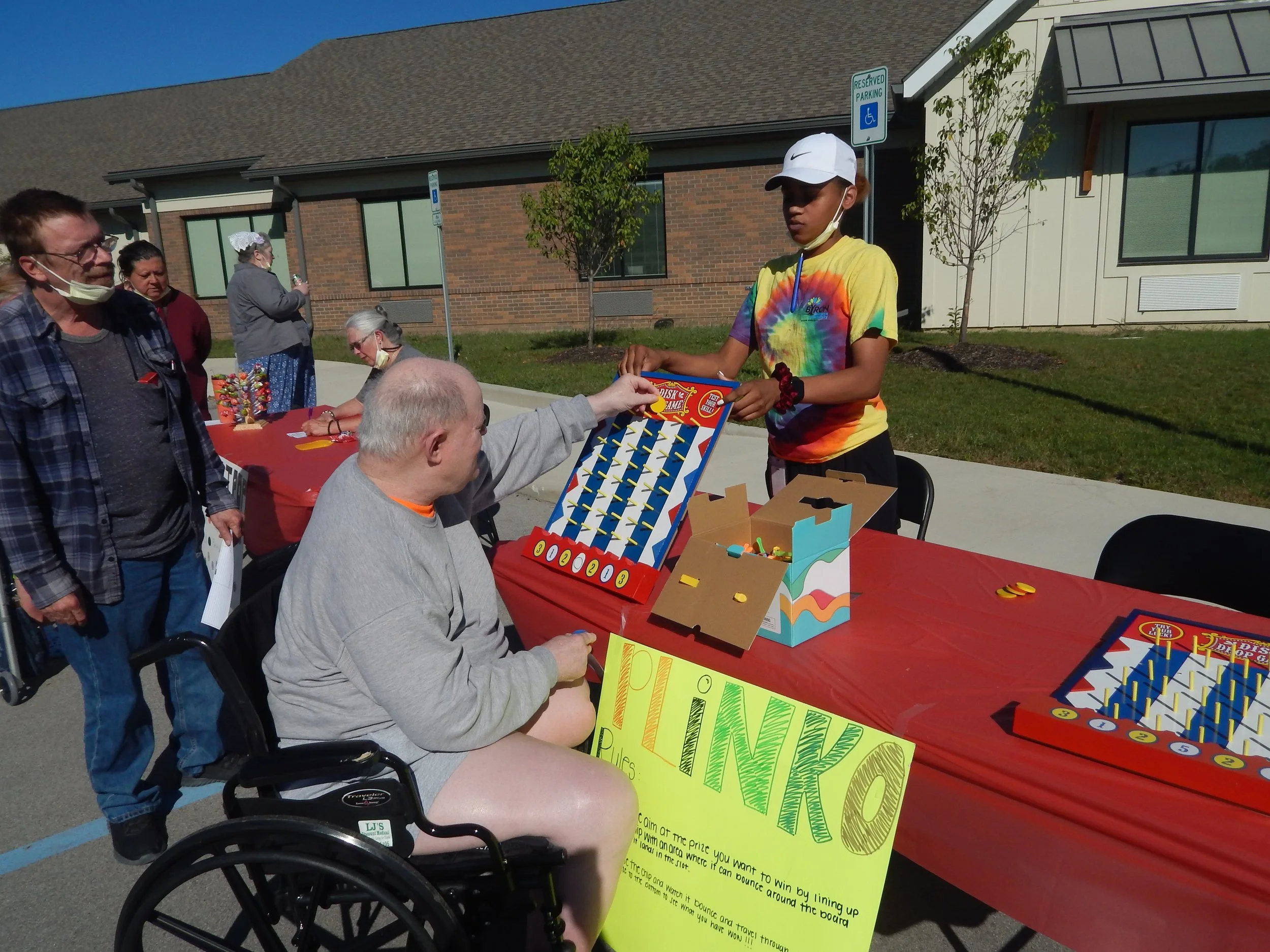 Young person in tie-dye t-shirt and white cap playing plinko game at outdoor event, with older people watching and chatting at table with candy and signs.