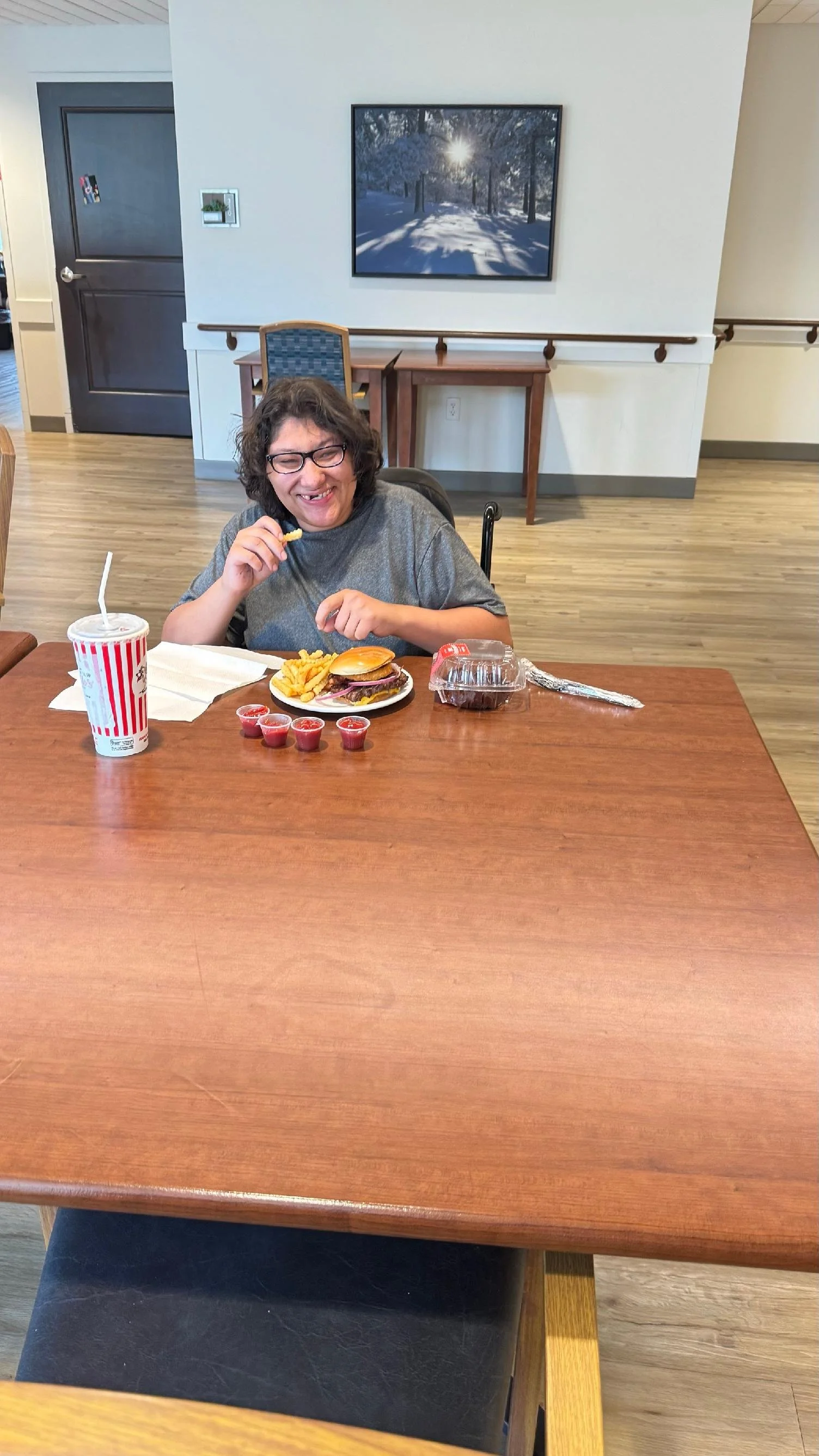 A smiling woman with glasses sitting at a dining table, holding a French fry, with a cheeseburger, French fries, small cups of sauce, a drink, and a container of chocolate pudding in front of her.
