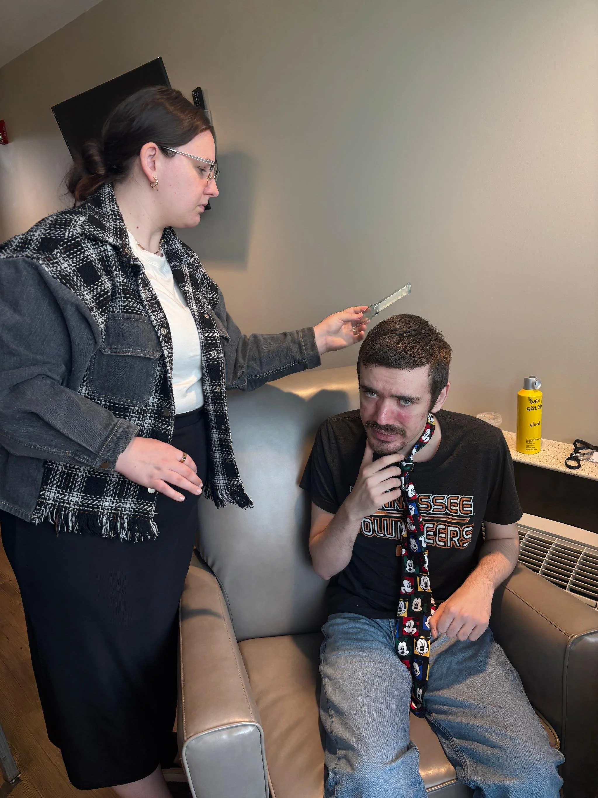 A woman with glasses and a patterned black and white jacket holding a hairbrush, appears to be styling or examining the hair of a man with dark hair, a beard, and a mustache, who is sitting on a beige couch. The man is wearing a black T-shirt with an