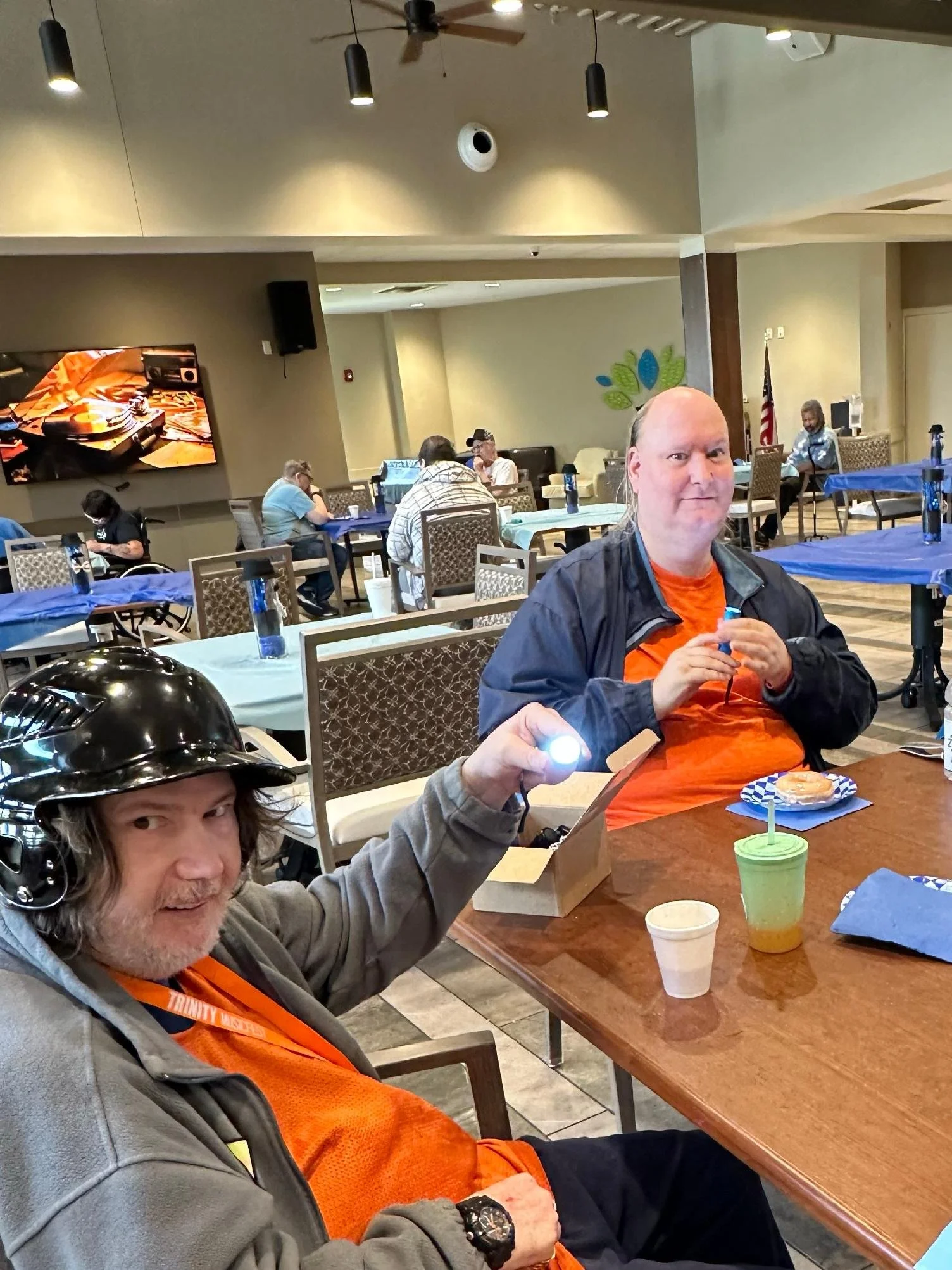 Two men in orange shirts are sitting at a table in a restaurant or cafe. One man is holding a small box and a green cup with a straw, and the other is holding a flashlight. There are other people in the background, some using laptops and phones, with