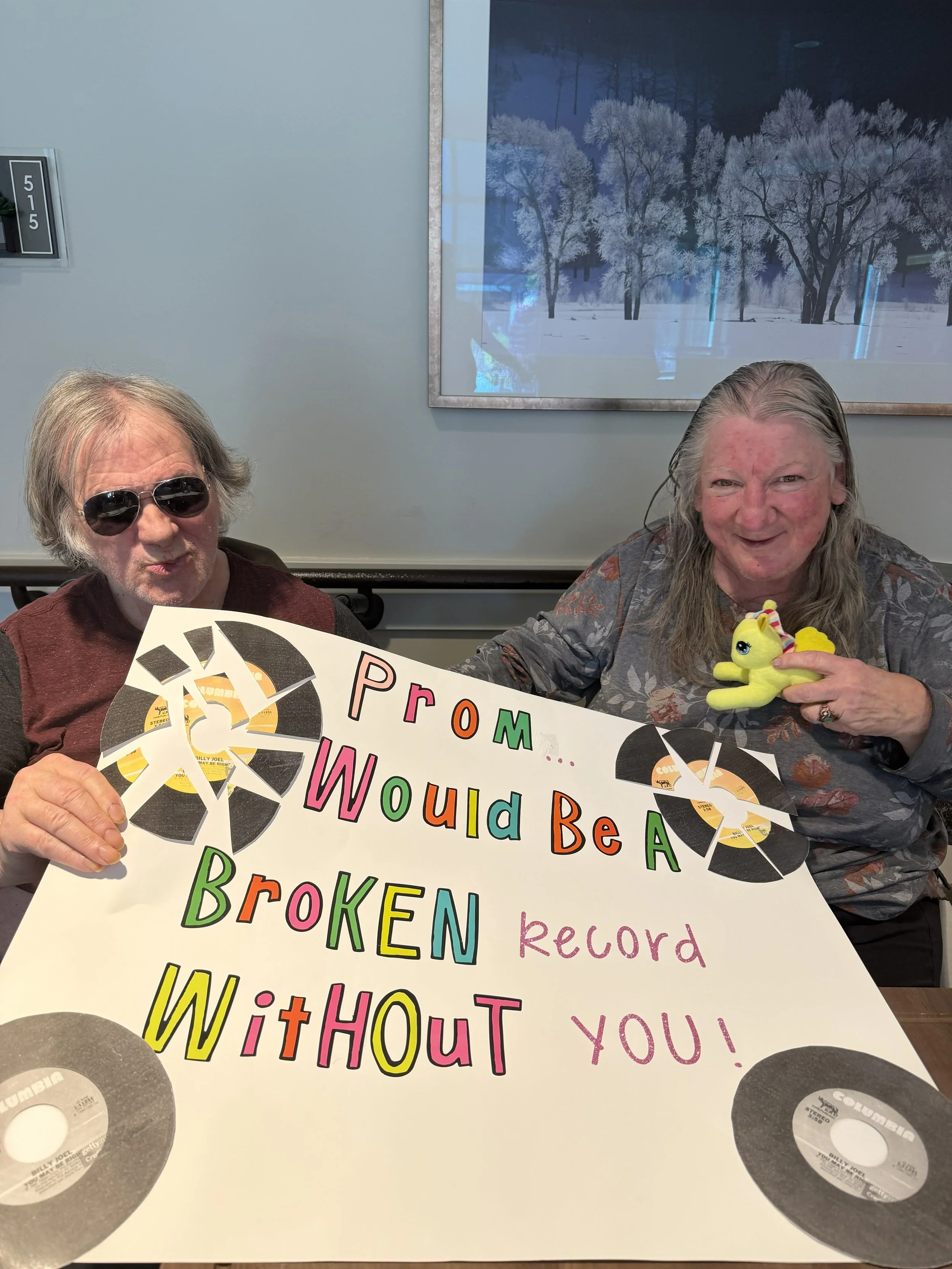 Two people holding a colorful protest sign with music records images attached, and smiling at the camera. The sign reads: 'PROMISE I WOULD BE A BROKEN record without you!'. One person holds a yellow plush toy.