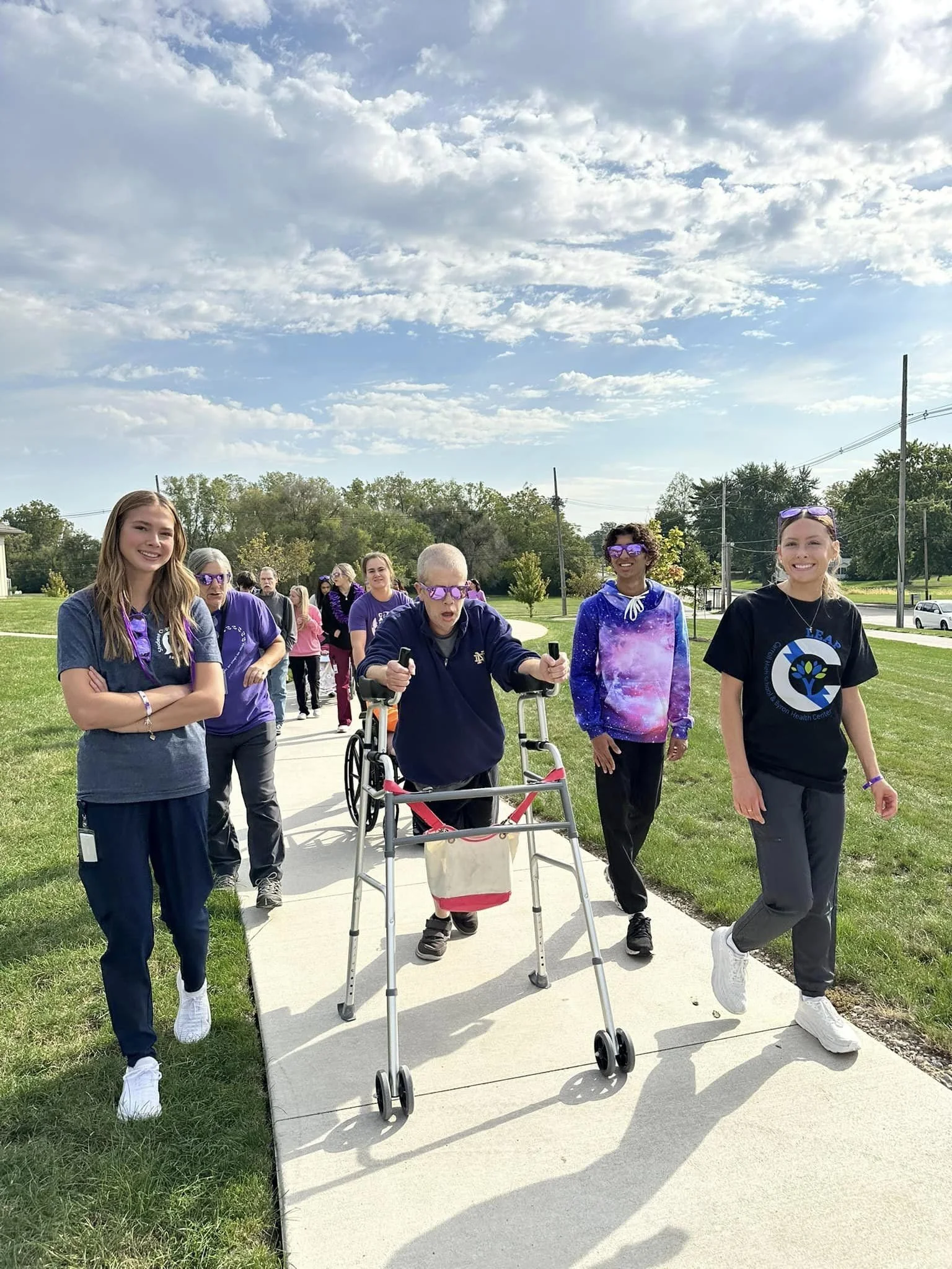 A group of young people walking outdoors on a sidewalk, with a man using a walker in the center. The sky is partly cloudy, and there are trees and utility poles in the background.