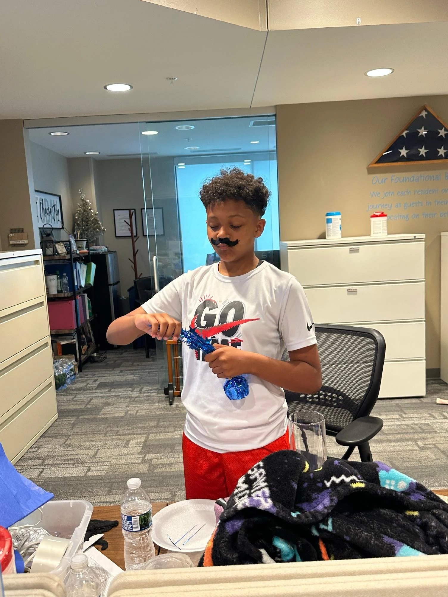 A young boy with curly hair and a fake mustache is standing in an indoor setting, holding a blue and red object. He is wearing a white T-shirt with a graphic design and red shorts. The room has filing cabinets, framed pictures, a small Christmas tree