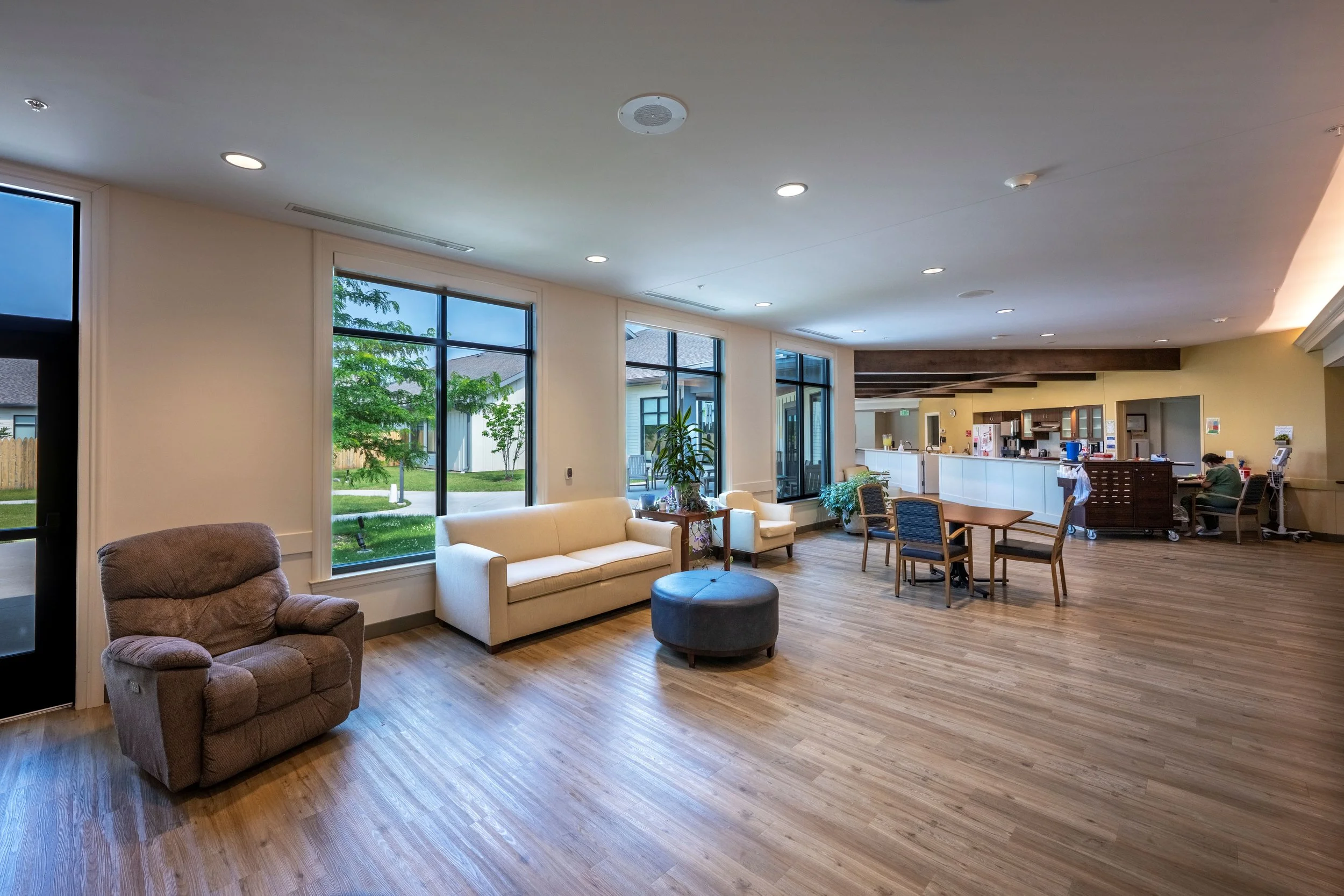 A spacious, well-lit common area with large windows showing greenery outside. The room is furnished with a brown armchair, cream-colored sofas, a round ottoman, and a dining table with chairs. In the background, a counter area with a person sitting and working, a kitchenette, and kitchen appliances are visible.