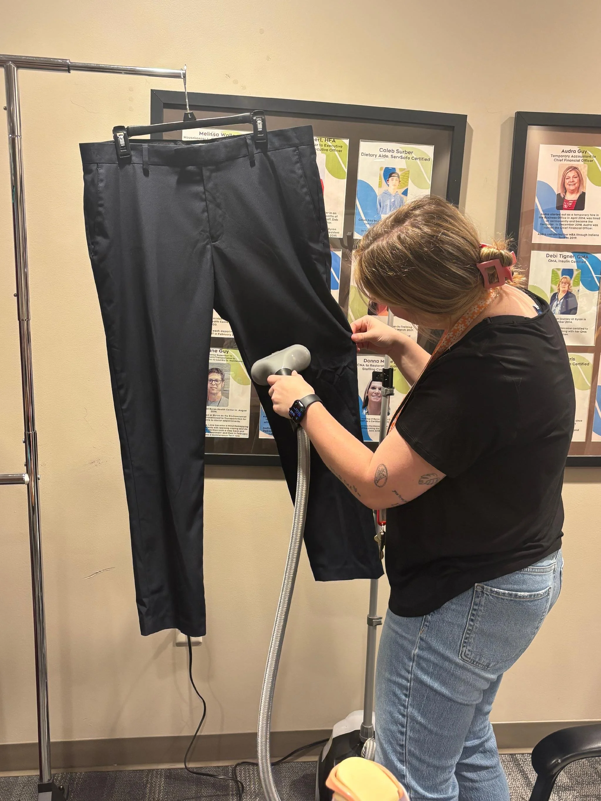 A woman with short brown hair and tattoos on her arm is using a handheld device on a dark pair of pants hanging on a clothing rack. The pants are on a hanger, and the woman is leaning forward, focused on the pants. There are posters with photos of pe