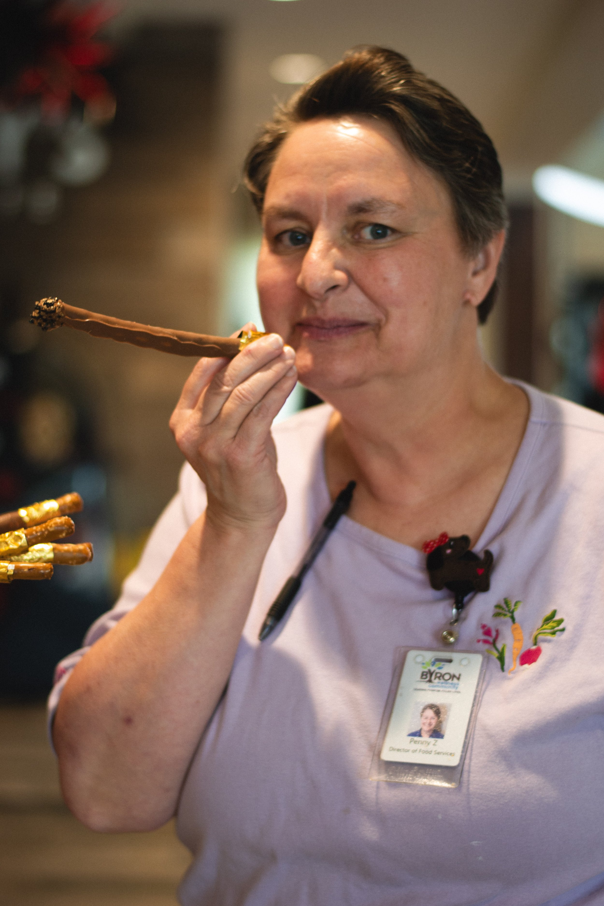 A woman with short dark hair holding a decorated drumstick near her face, wearing a pink uniform with a name badge and a lanyard, standing in a well-lit indoor environment.