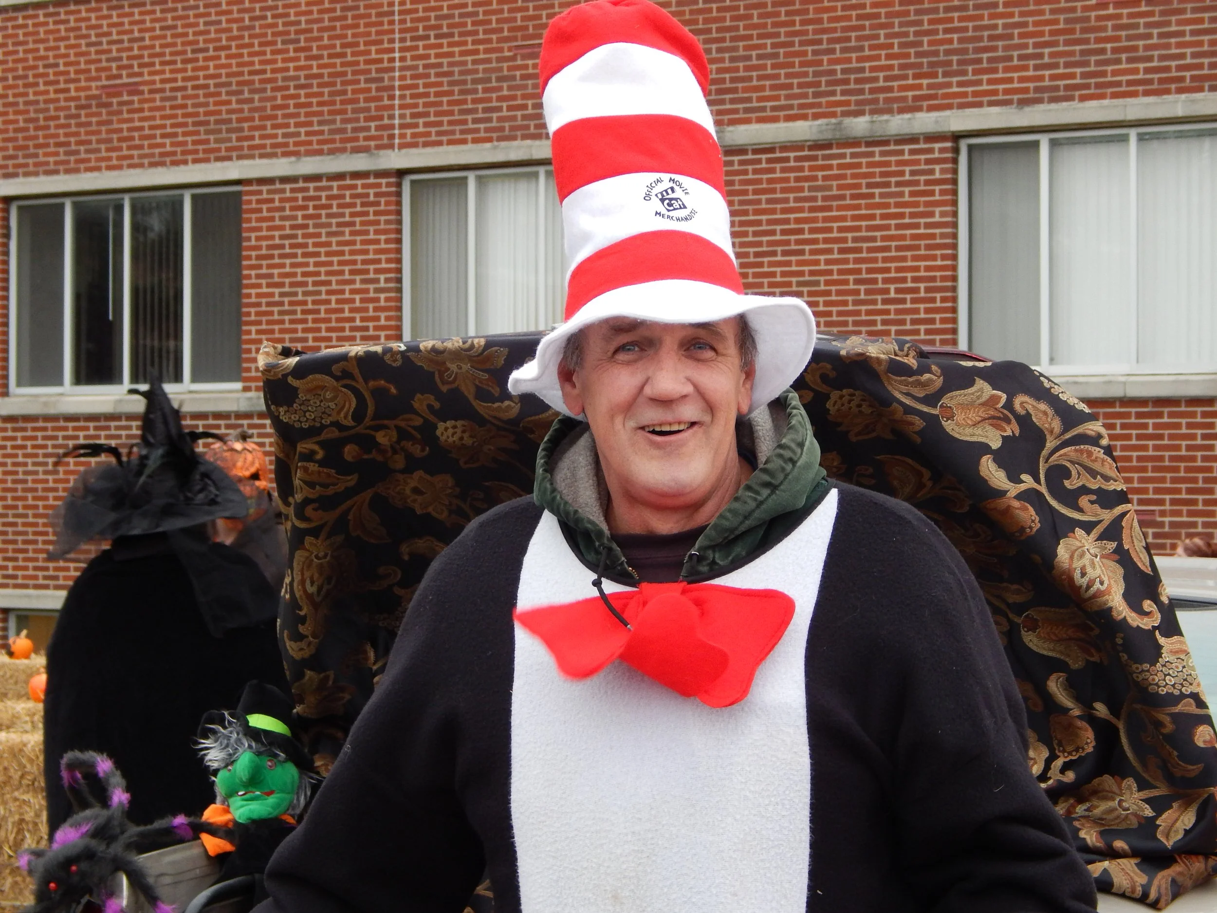 A man dressed in a Dr. Seuss-themed costume wearing a tall red and white striped hat with a logo, smiling outdoors in front of a brick building with windows, during a festive event with Halloween decorations and pumpkins.