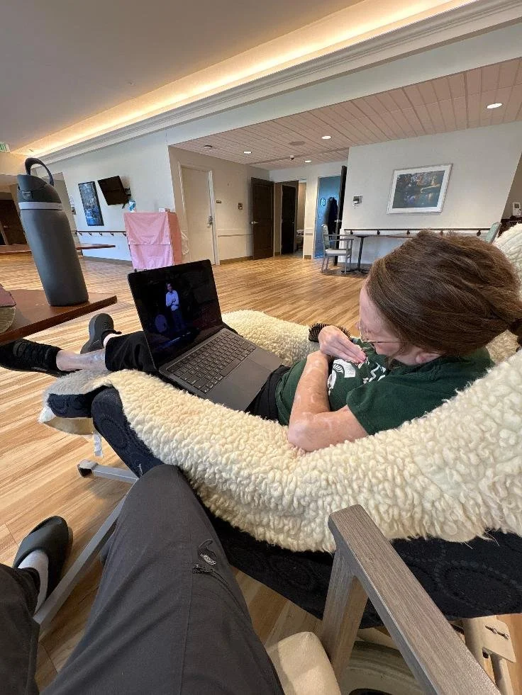An elderly woman lying on a lounge chair watching a video call on her laptop, in a spacious waiting area or lounge with wooden floors and modern decor.