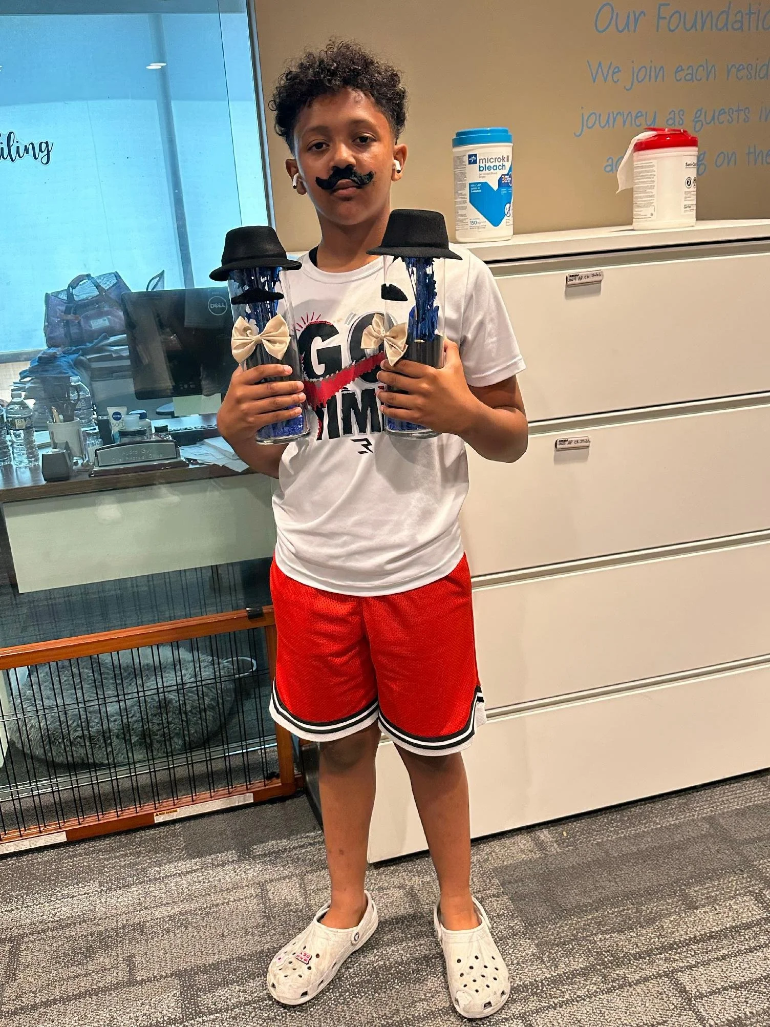 A young boy with a faux mustache is holding two jars with black hats and bowties decorated on them, in an indoor setting with filing cabinets and a glass window in the background.